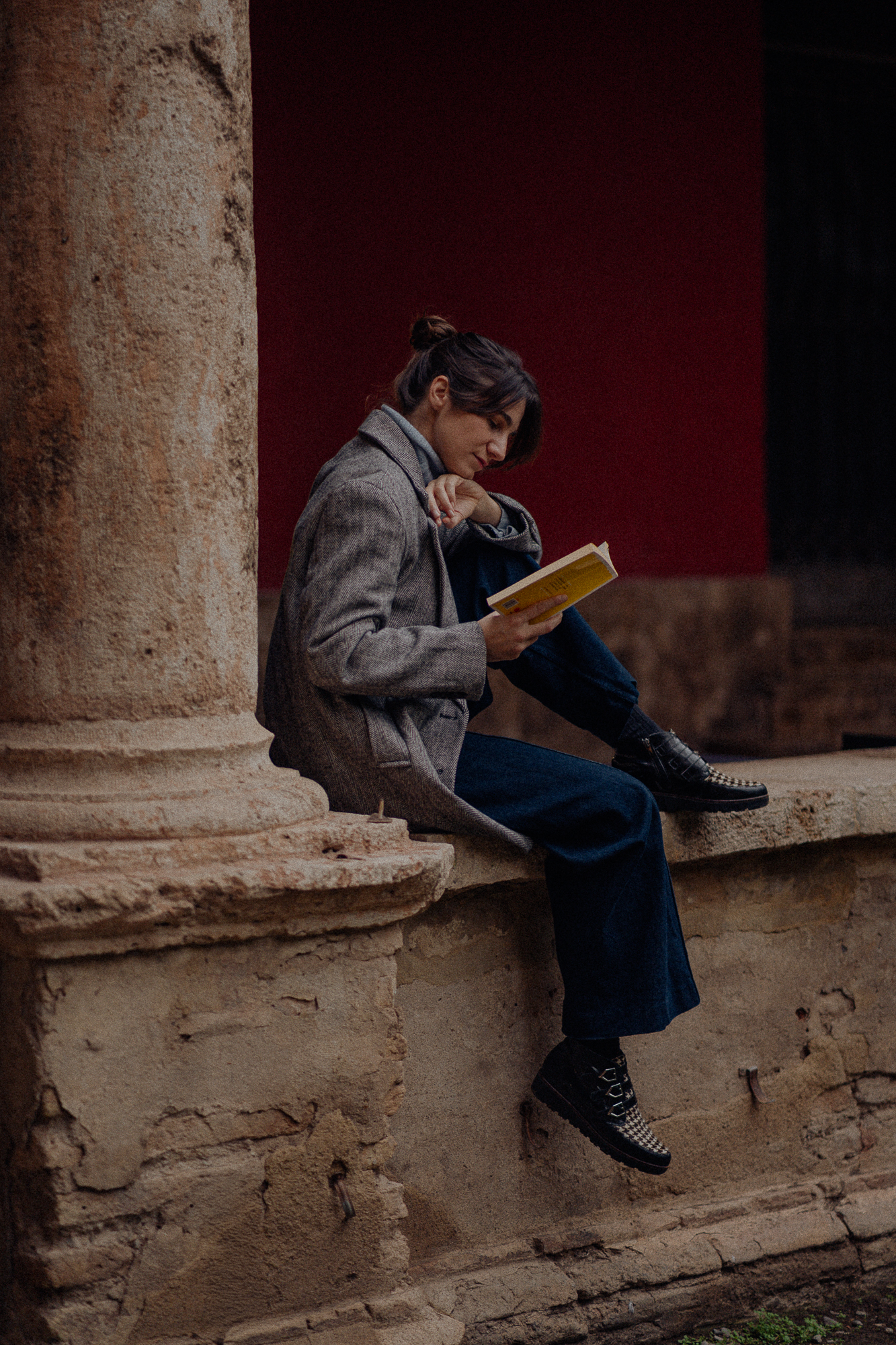 A woman sitting on stone steps at the base of a large pillar, reading a yellow book. She is dressed in a gray coat, blue pants, and black and white checked shoes. The background features dark red and black walls.