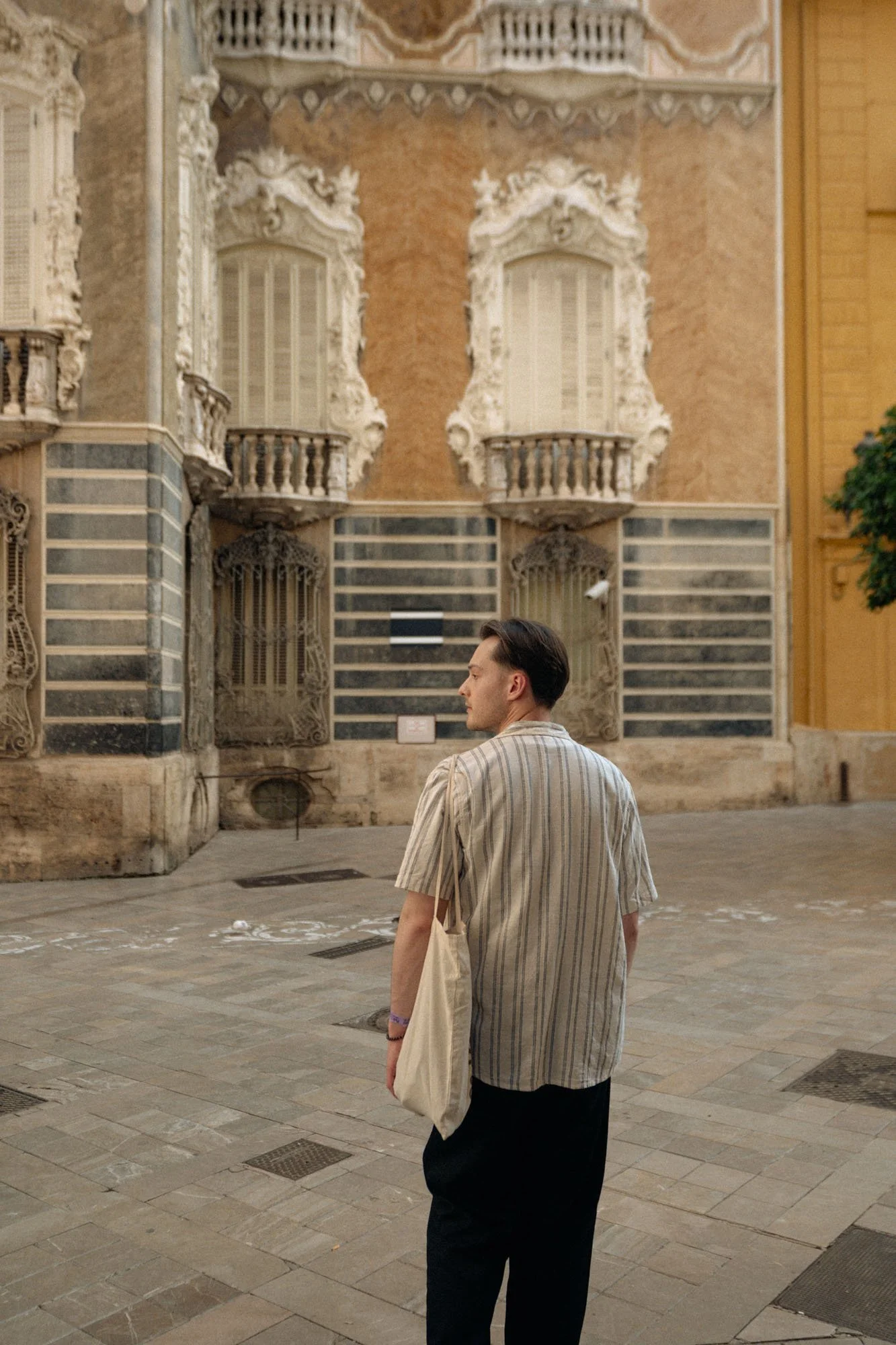 A young man with dark hair, wearing a beige striped shirt, black pants, and carrying a beige tote bag, stands outdoors in front of an ornate historical building with intricate architectural details and balconies.