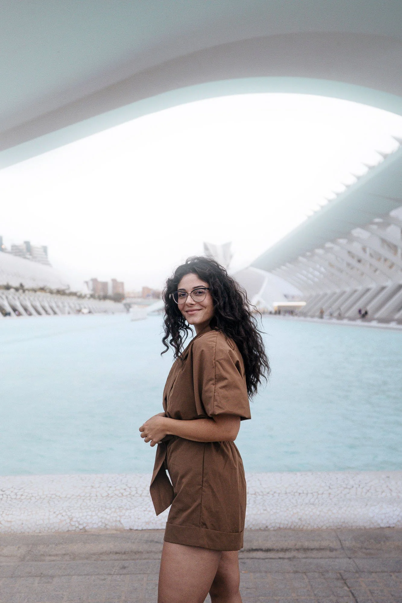 A woman stands outdoors near a water body with a modern architectural structure behind her, wearing glasses and a brown shirt with matching shorts.