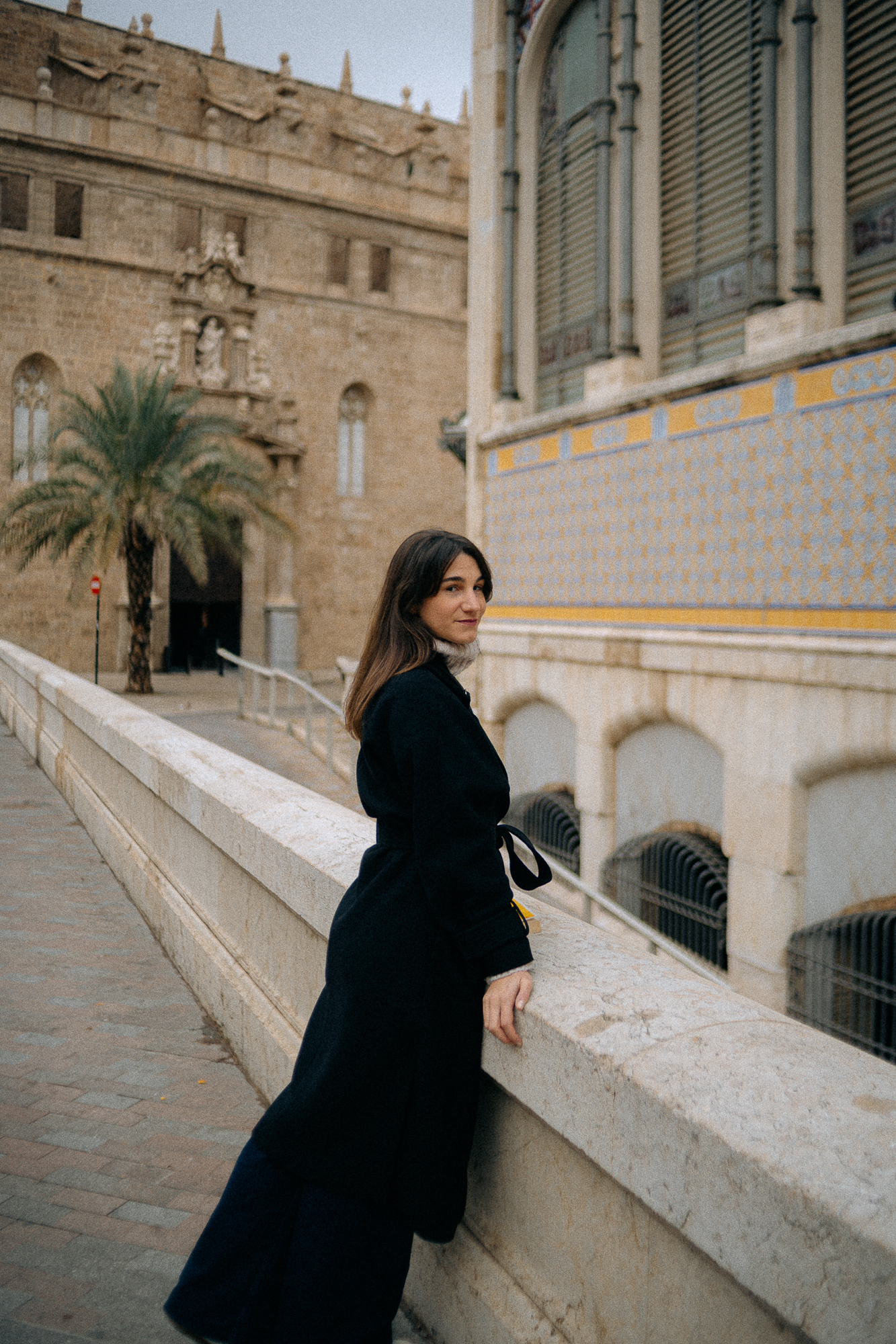 A woman with long dark hair, wearing a black coat, standing beside a stone railing, looking at the camera with a slight smile in front of a historic building with detailed architectural elements, arched windows, and blue and yellow decorative tiles.