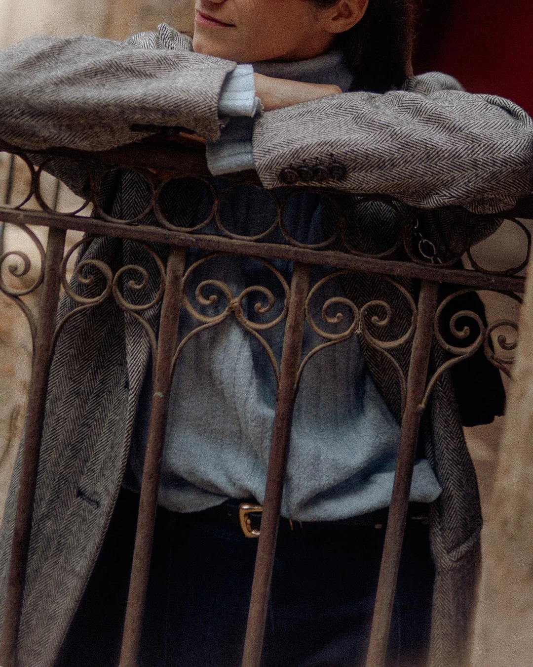 Person leaning on an ornate iron railing with curly patterns, wearing a gray plaid blazer and a gray shirt.