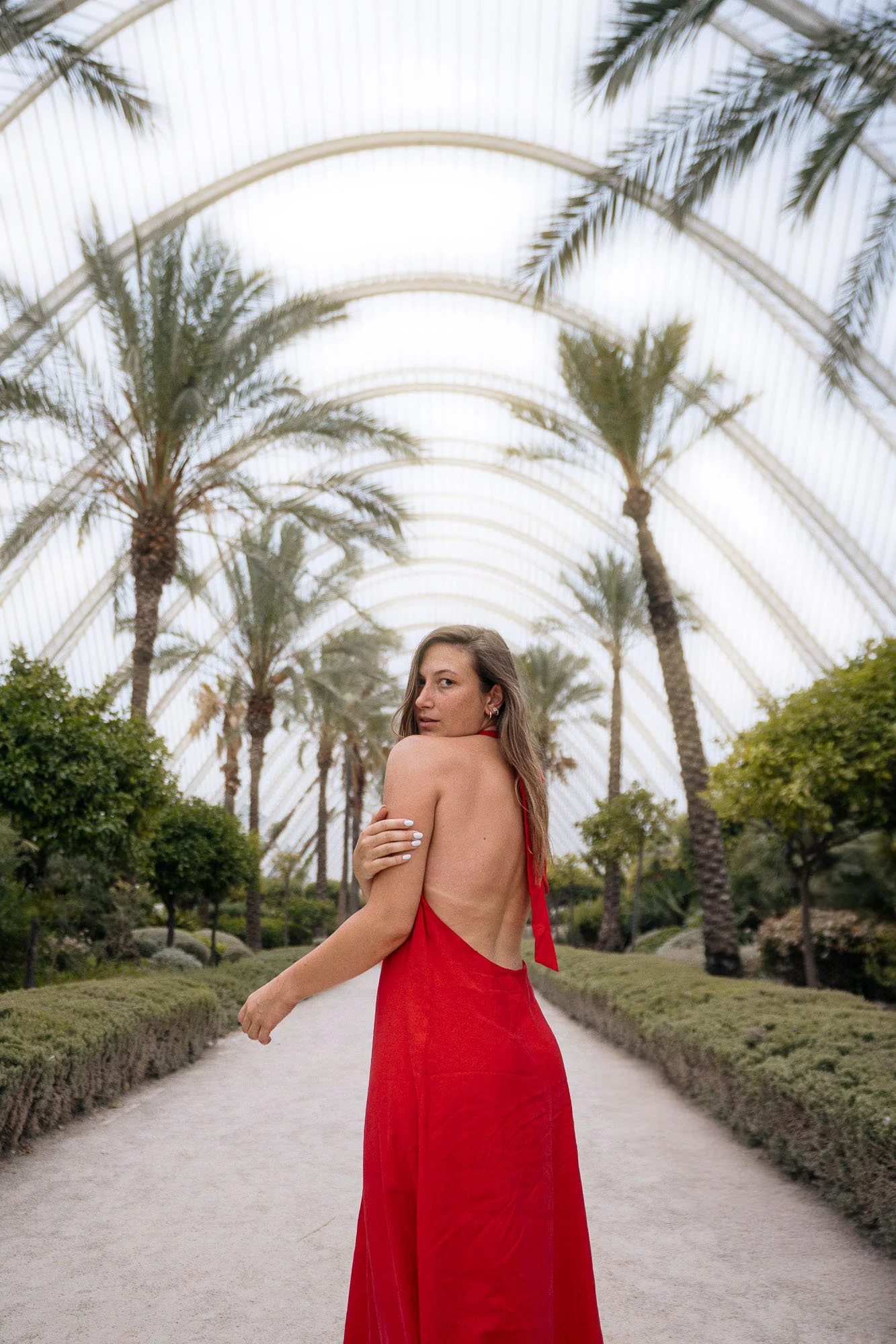 A woman in a red dress with an open back standing on a pathway lined with greenery and tall palm trees under a glass arch ceiling.