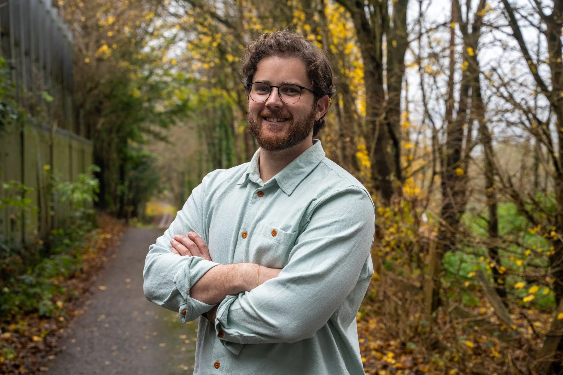 A smiling man with glasses and a beard, wearing a light-colored button-up shirt, stands with arms crossed on a fall day in a wooded area with trees and fallen leaves. Archie Lasseter, Archibald Lasseter