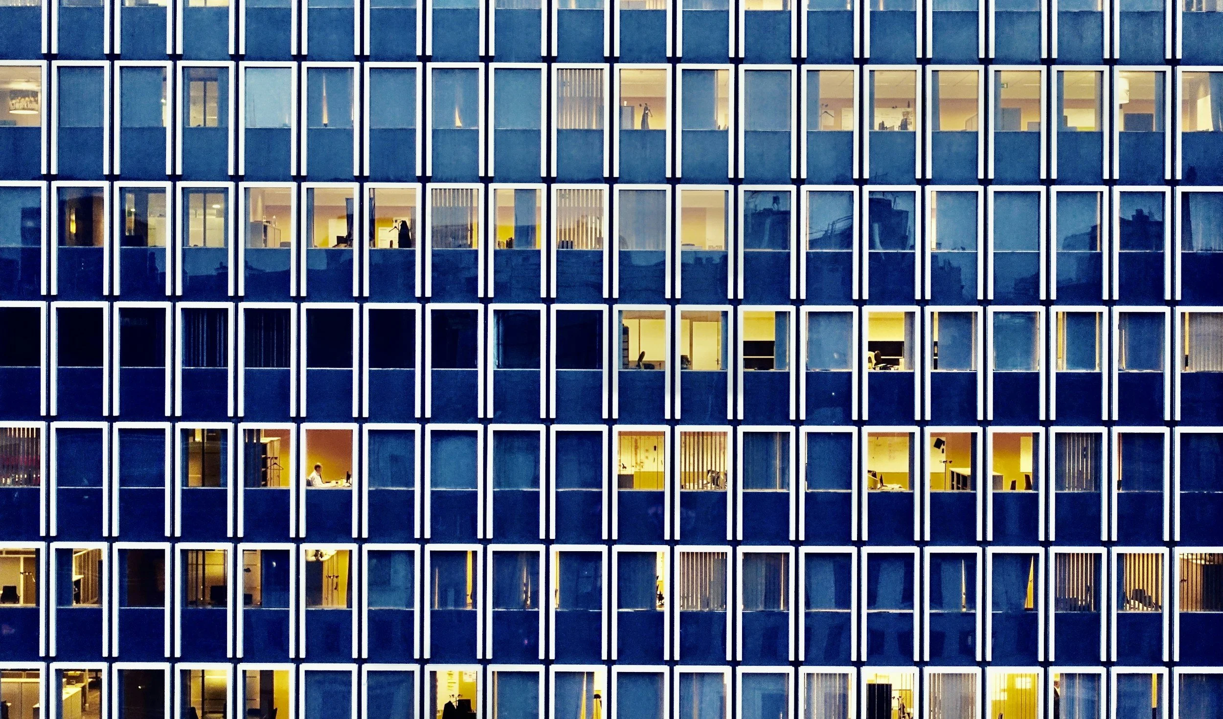 The image shows the exterior of a modern office building at dusk, with illuminated office spaces visible through the large grid-patterned windows. Some office workers are seen working inside, and the overall scene suggests a busy urban environment.