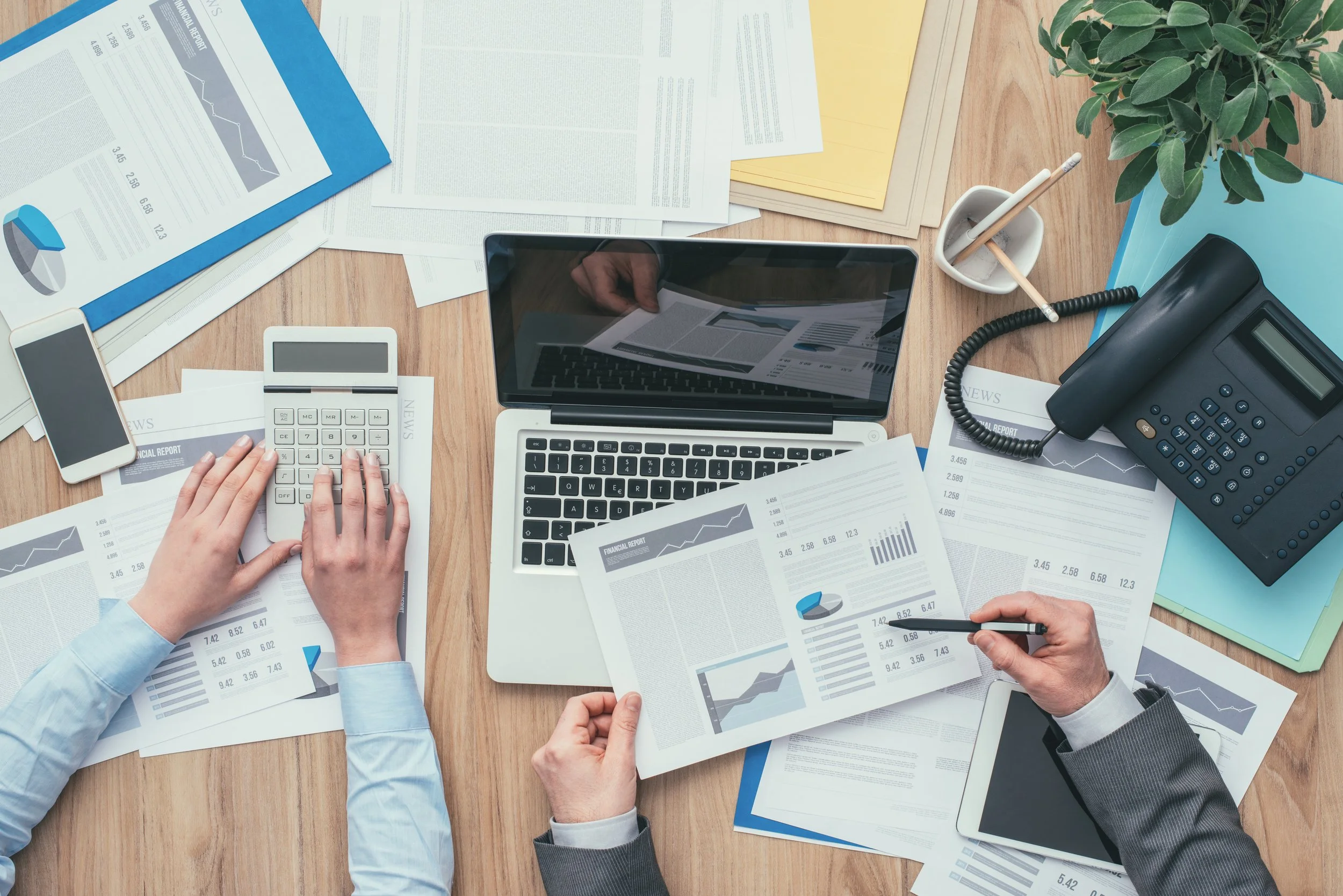 A cluttered wooden desk with scattered business documents, financial reports, charts, and graphs. Several laptops, a smartphone, calculator, landline phone, and writing utensils are also on the desk, with hands of multiple people working and pointing at the papers.