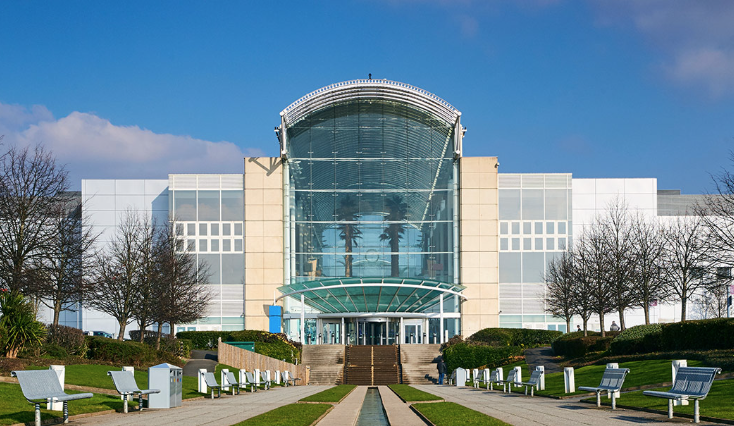 Modern building with large glass front entrance, surrounded by benches and trees, under a blue sky.