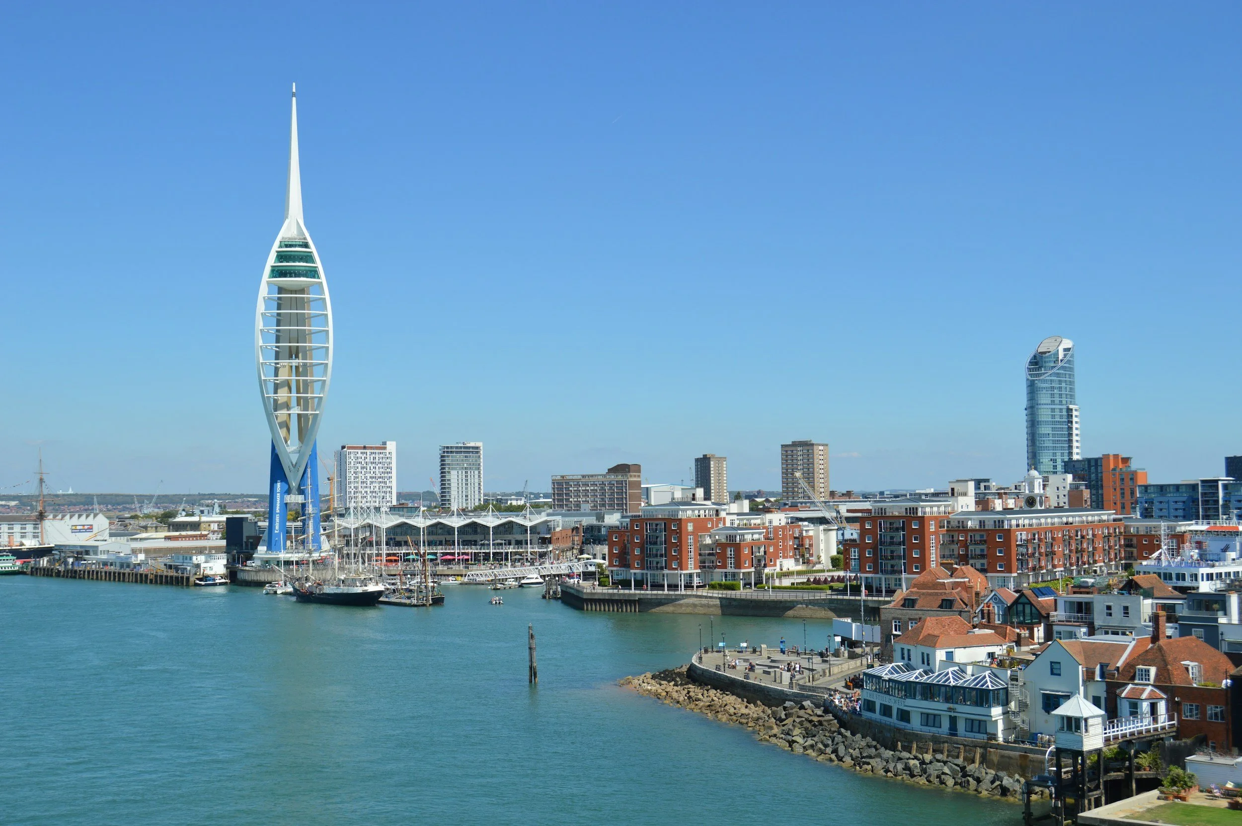 A city skyline with a prominent modern, sail-shaped building, boats docked at a marina, and colorful low-rise buildings along the waterfront under a clear blue sky.