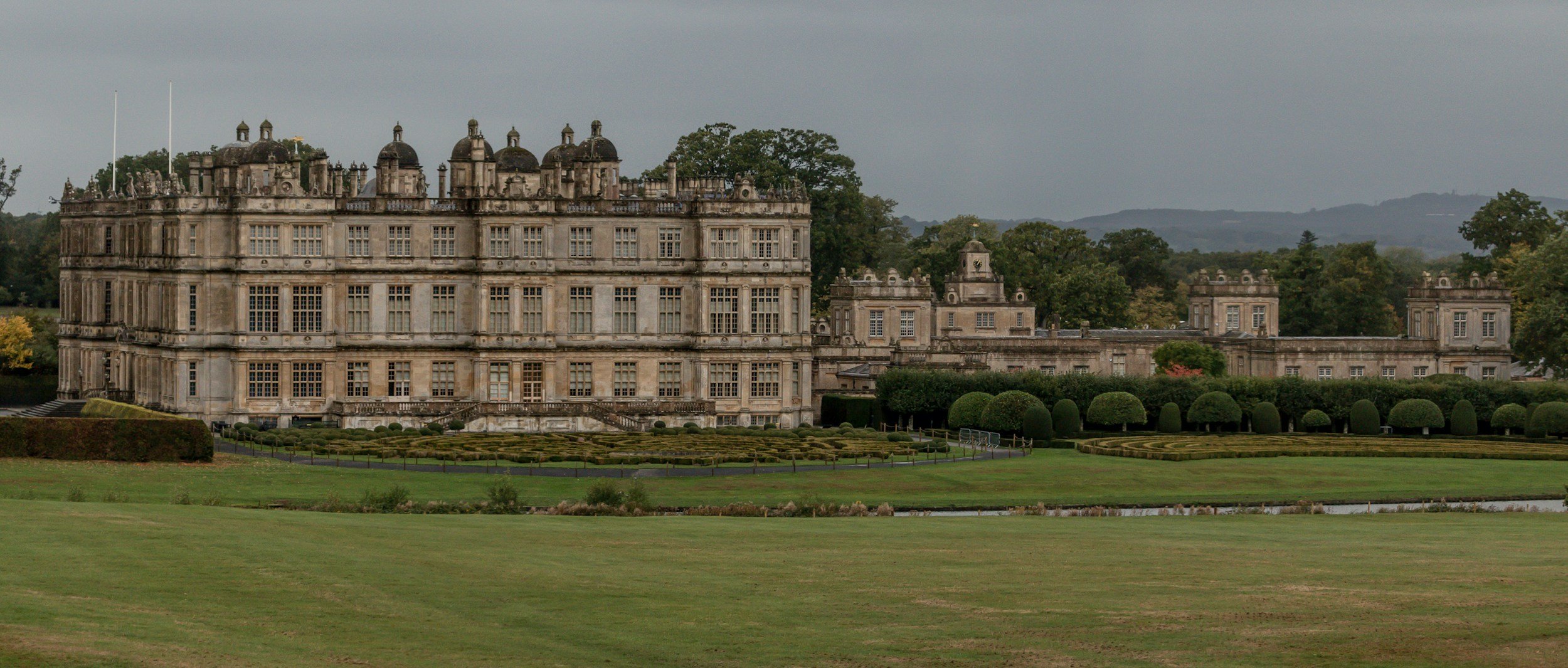 A large historic castle or mansion with multiple levels and detailed stone architecture, surrounded by manicured gardens and green lawns, with trees and hills in the background under a cloudy sky.