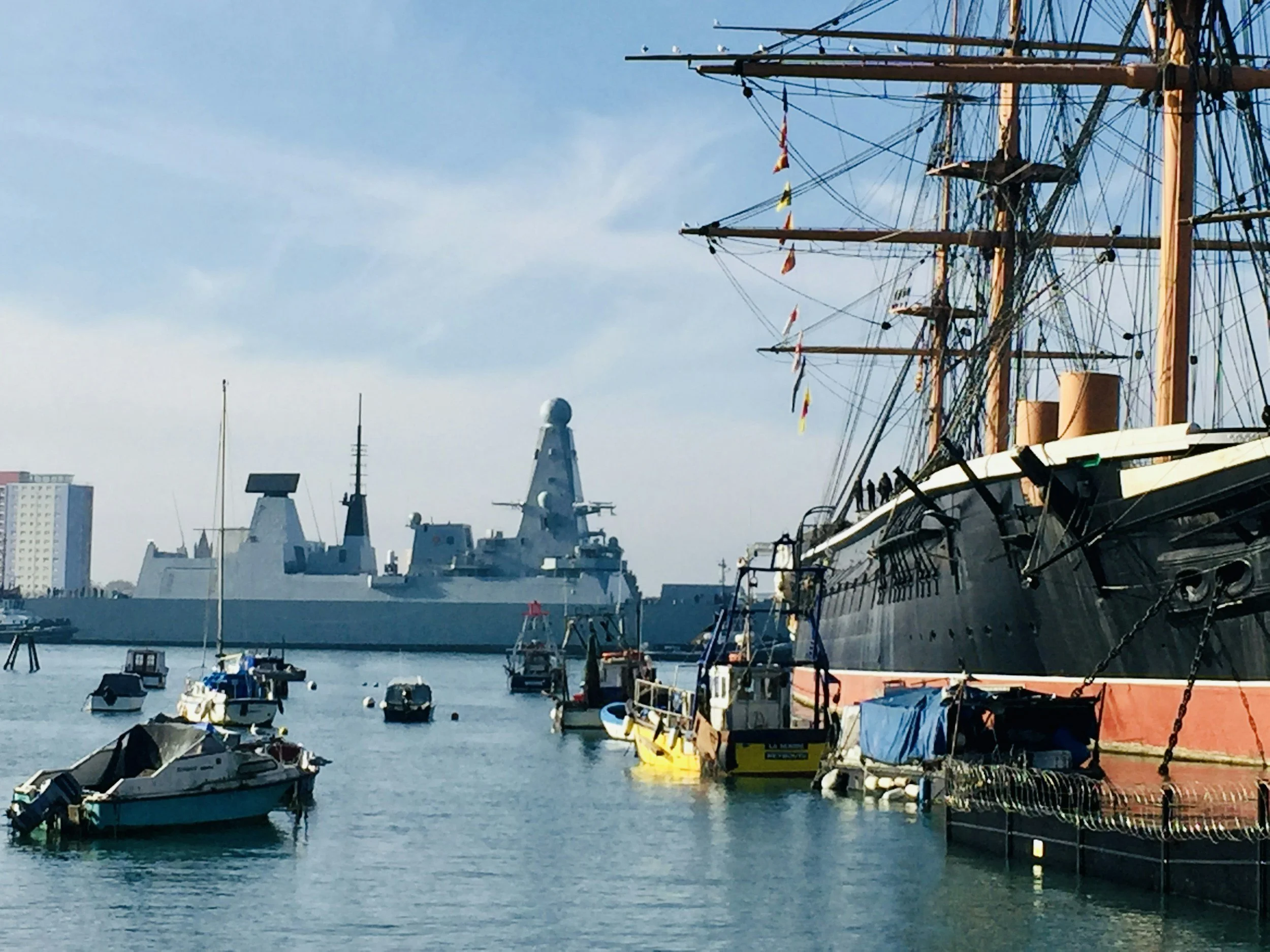 A harbor with various boats docked, including a large wooden sailing ship with tall masts and flags, and a modern navy ship in the background, under a blue sky.