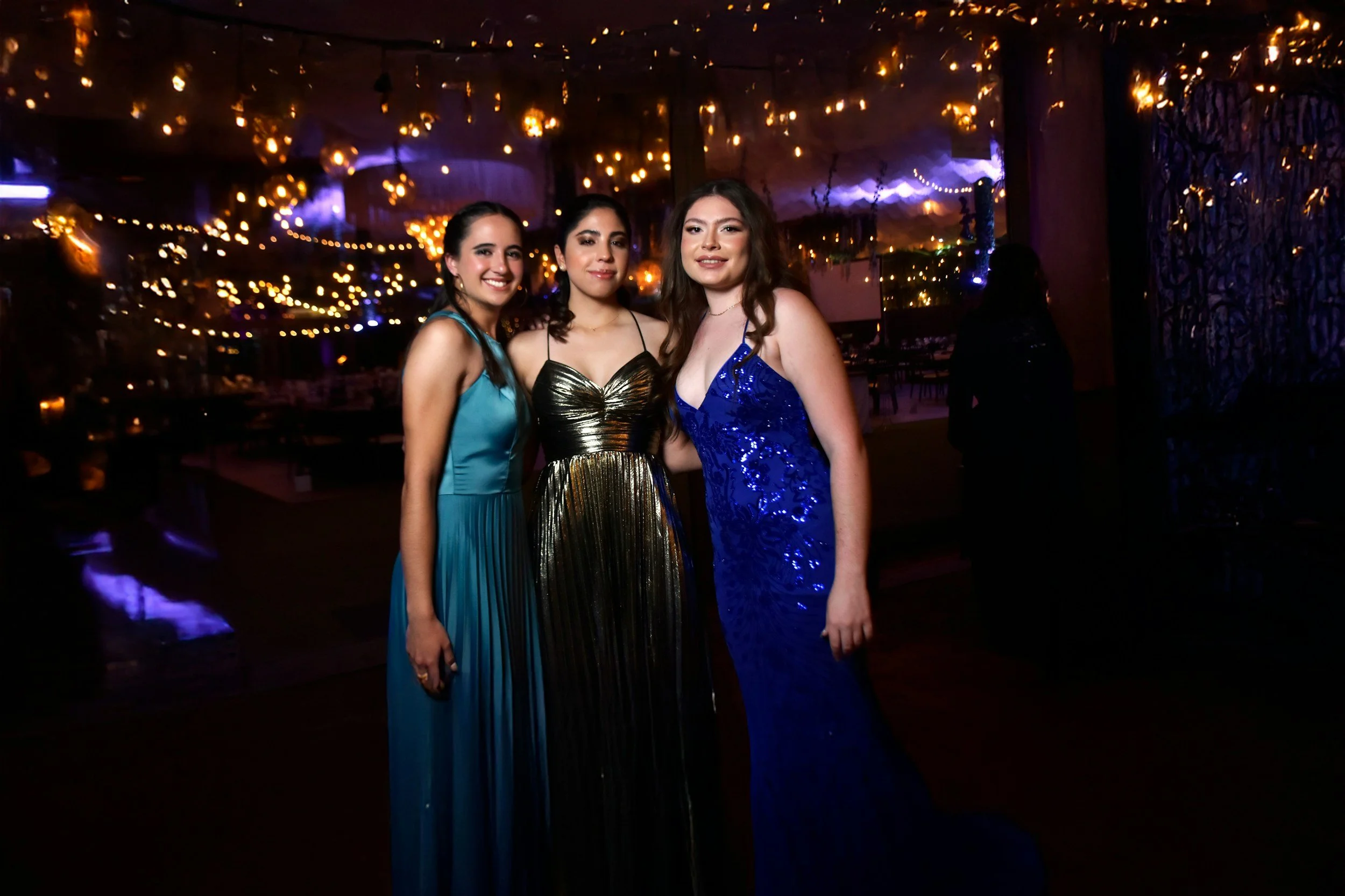Three young women in formal dresses posing for a photo at an indoor event with string lights and decorative lighting on the ceiling.