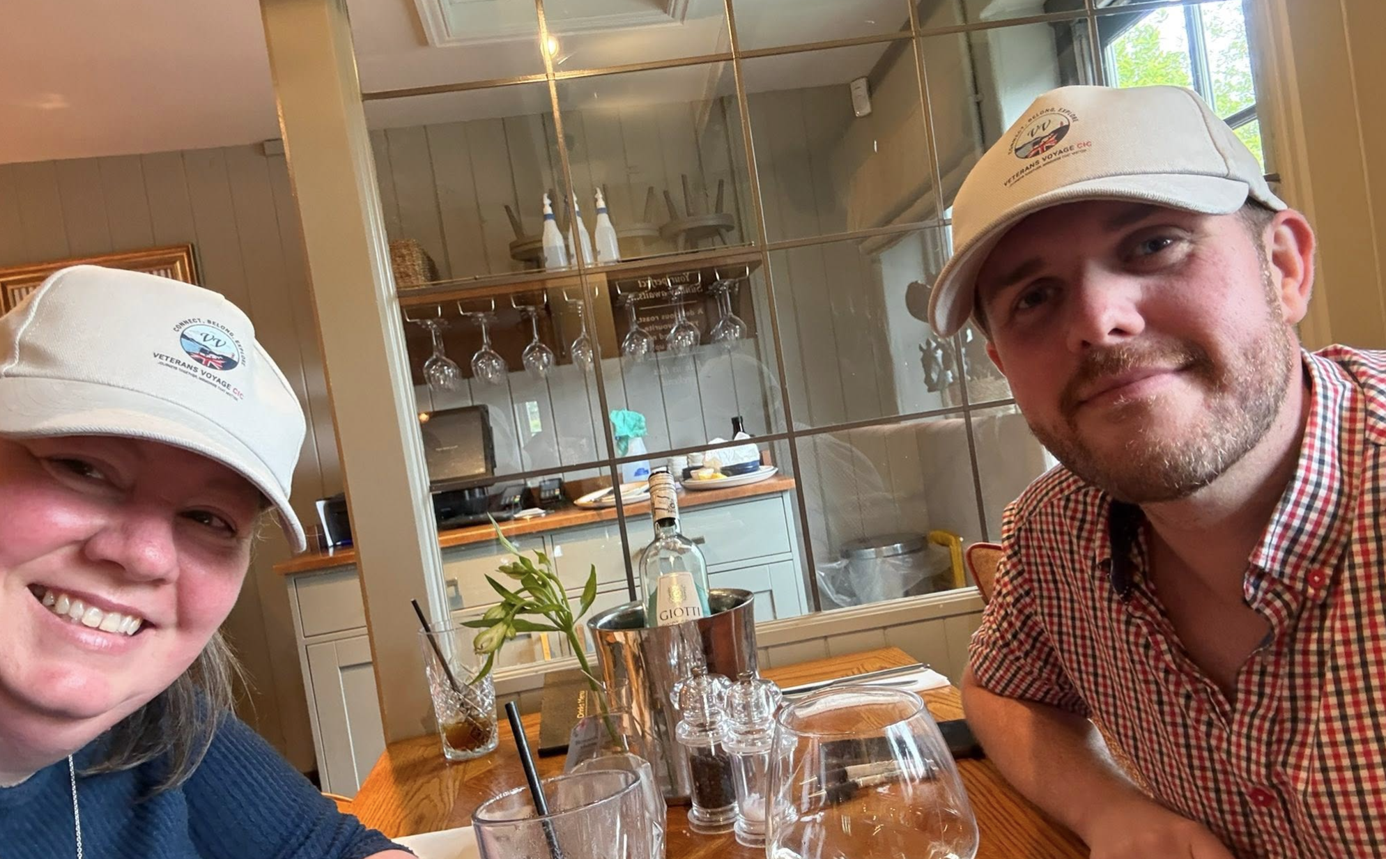 Two people smiling at a restaurant table, wearing Veterans Voyage hats, with drinks and a small plant on the table, and a glass wall in the background.