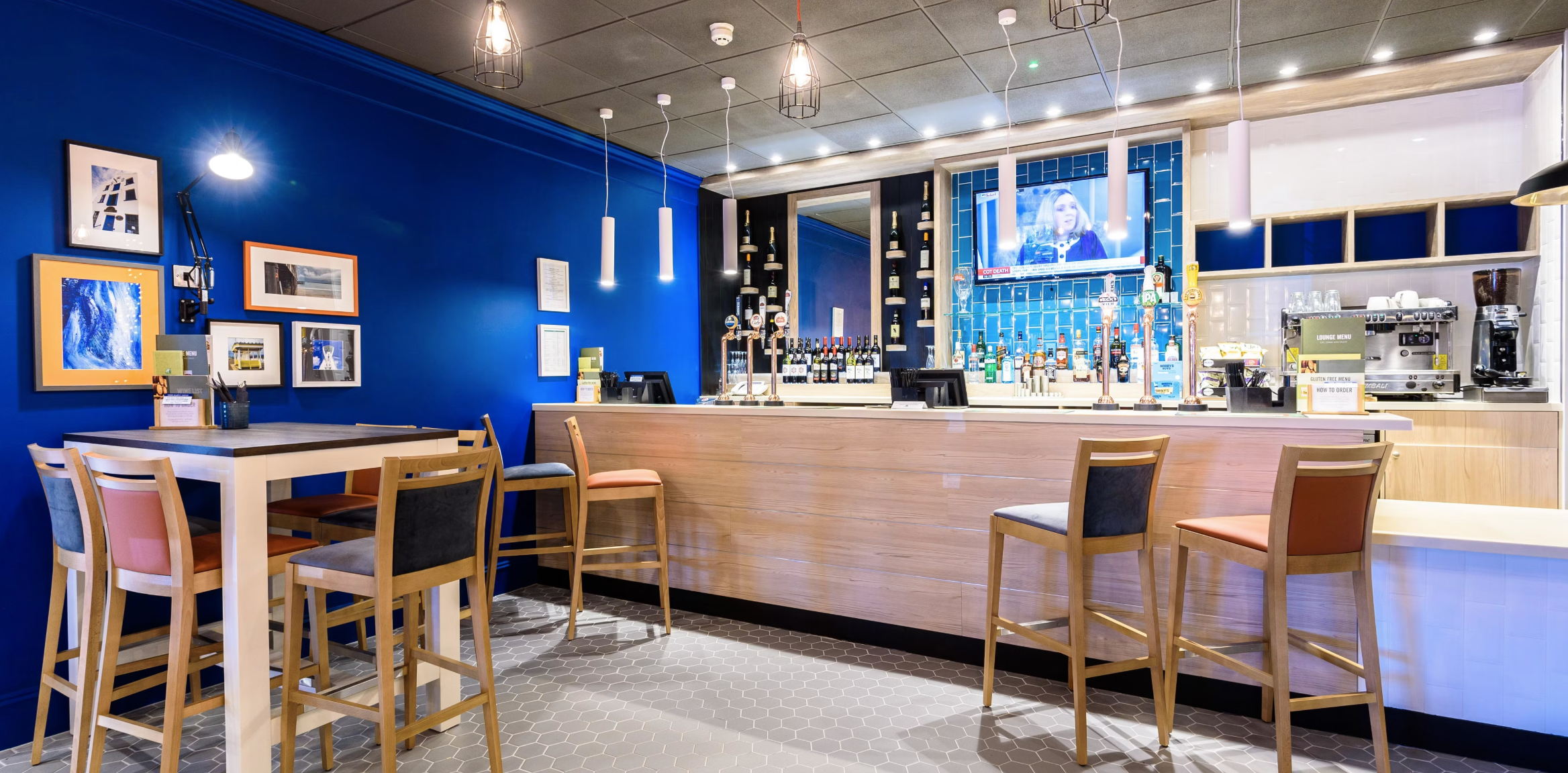 Interior of a modern bar with a blue accent wall, framed artwork, and a wooden bar counter with various bottles of alcohol and a television mounted above.