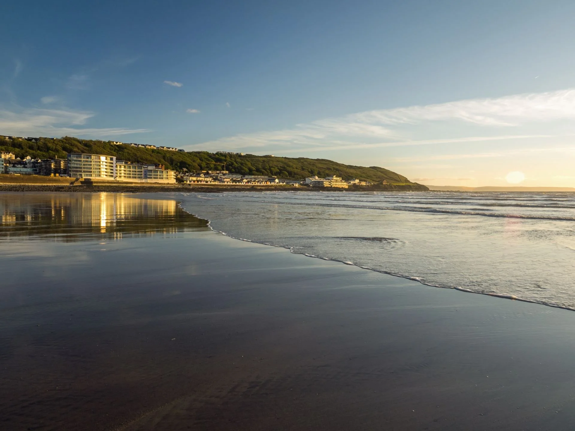 A beach at sunset with waves gently crashing on the shore, buildings and a green hillside in the background.