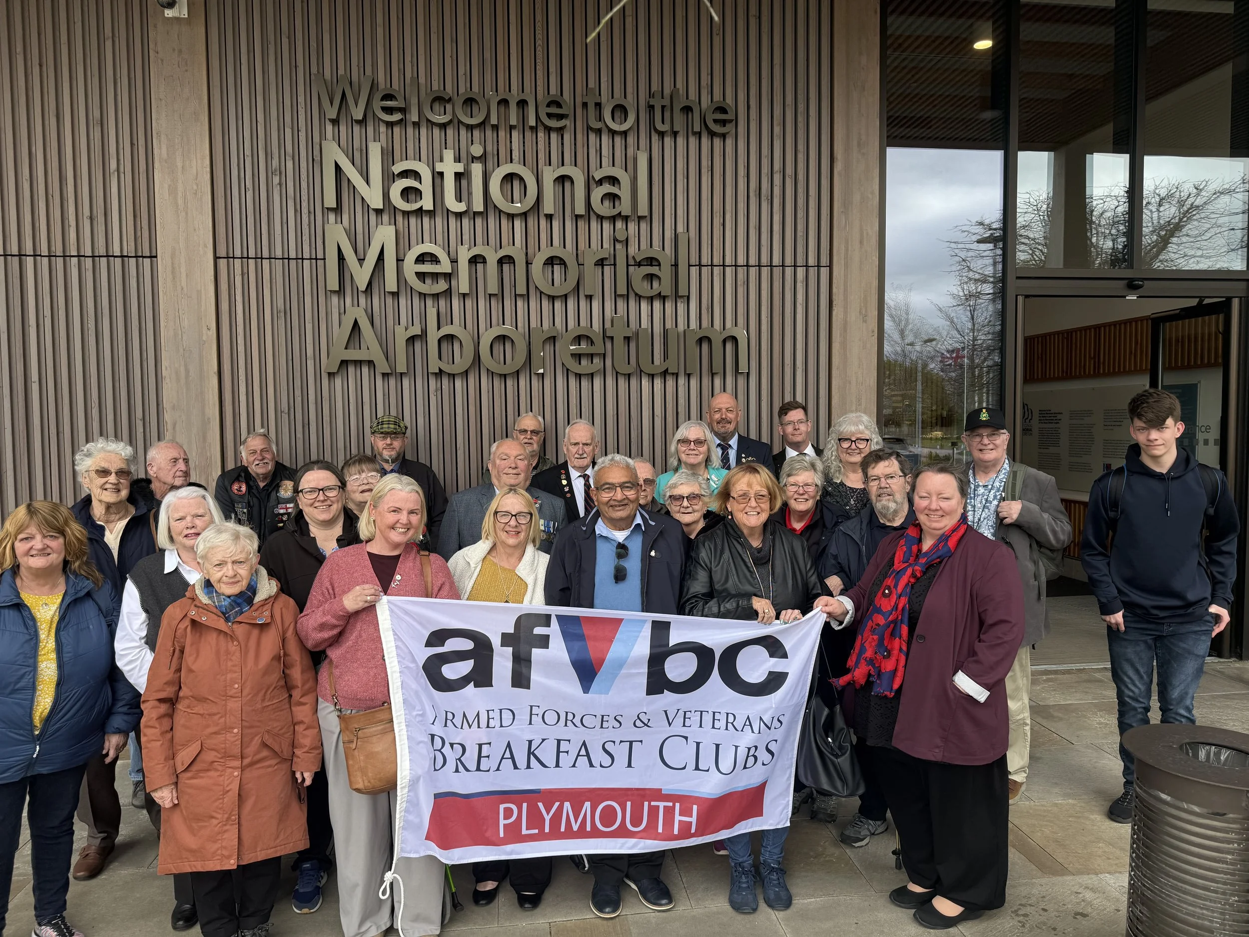 Group of veterans and community members holding a large banner in front of the National Memorial Arboretum, with a building displaying a sign that reads "Welcome to the National Memorial Arboretum."
