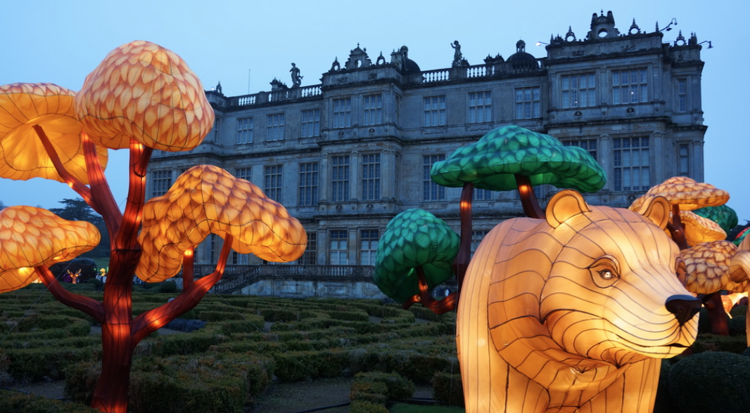 Illuminated lantern sculptures of trees and a lion cub in front of a historic building at dusk.
