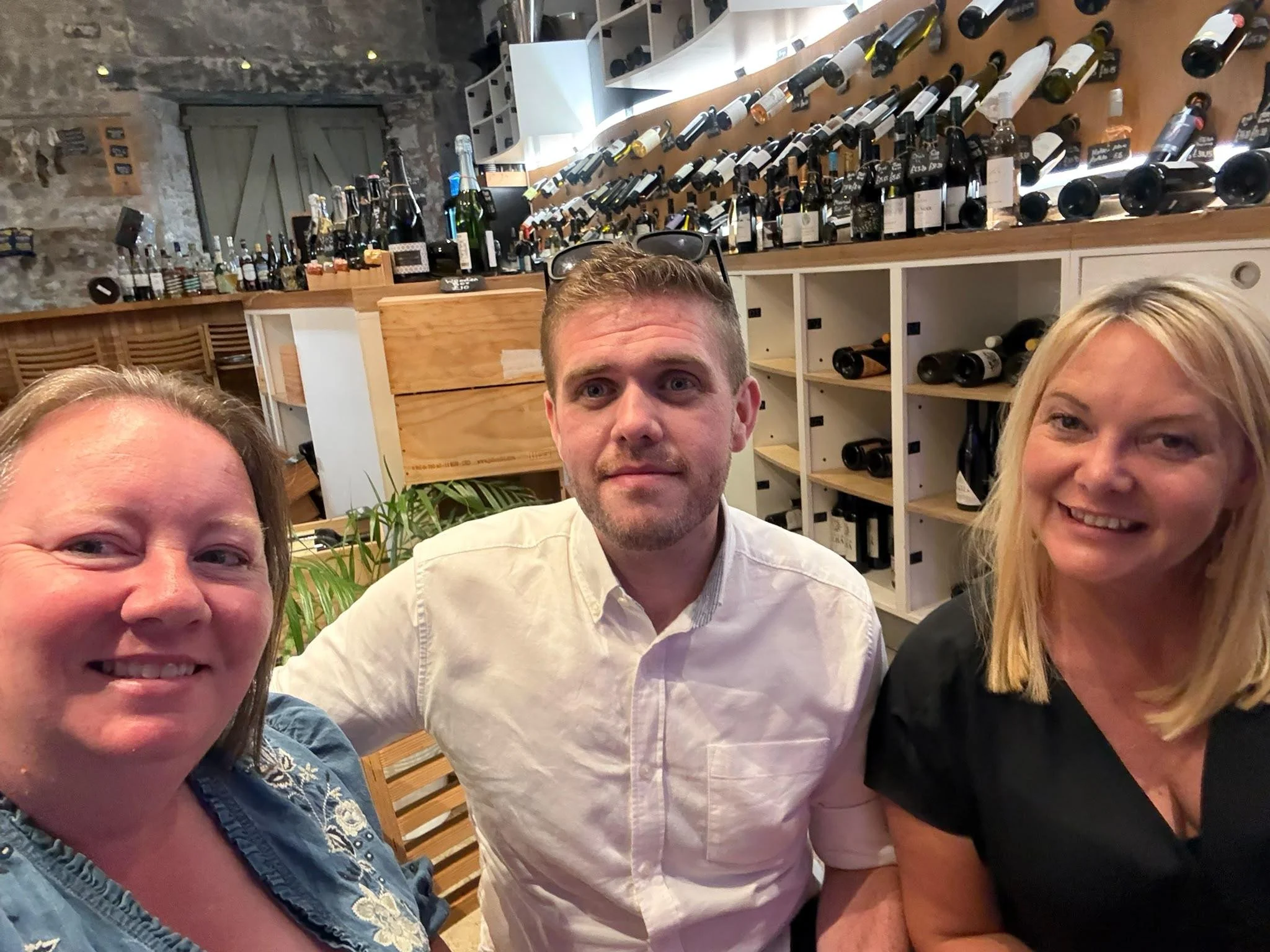 Four people taking a selfie in a wine cellar or wine bar with wine bottles stored on shelves behind them.