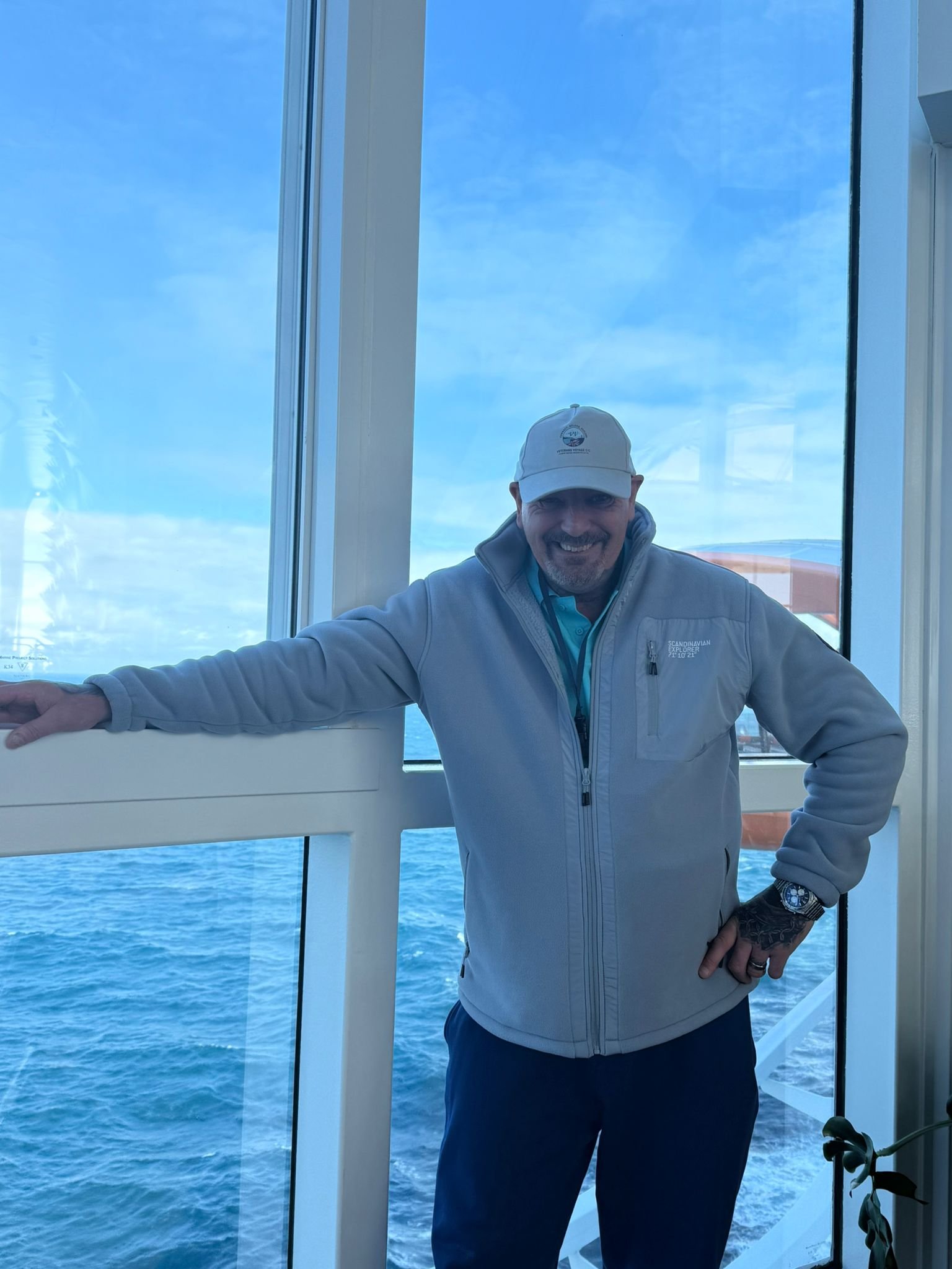 A smiling man in a light gray jacket and white cap stands near a window overlooking the ocean. The sky is clear with some clouds, and the water is blue.