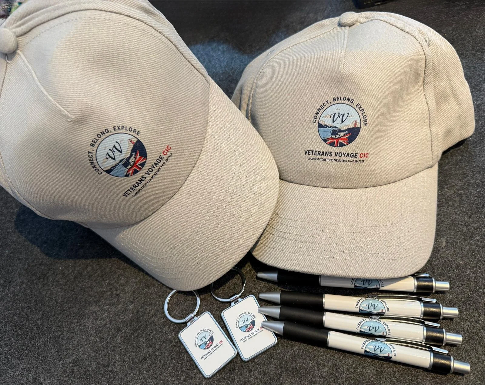 Three beige baseball caps with a circular logo featuring a boat and the words "Connect. Belong. Explore." and "Veterans Voyage CIC" along with some pens and keychains with the same logo.
