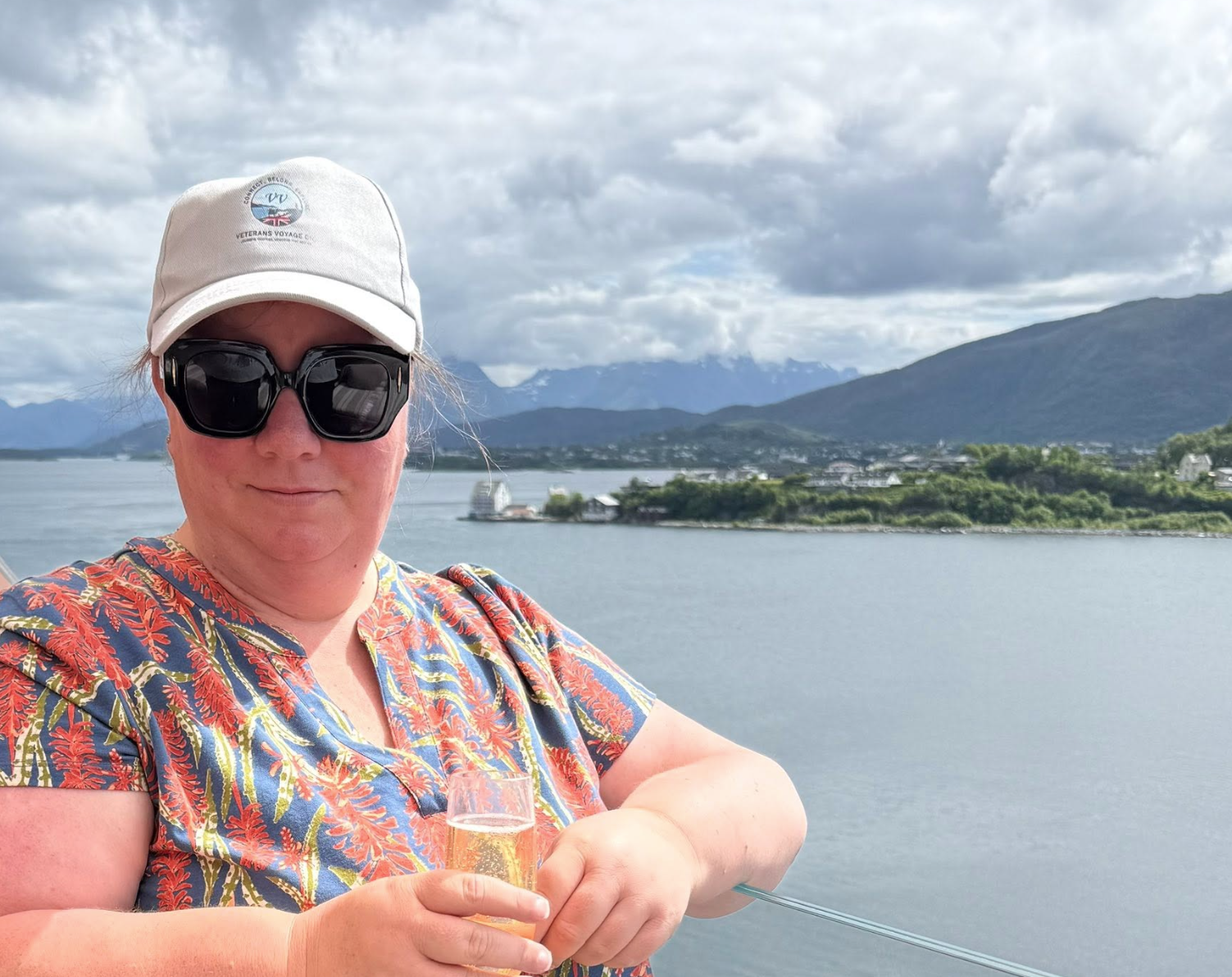 A woman wearing sunglasses, a white cap, and a floral shirt is holding a glass of champagne on a boat, with a scenic lake, mountains, and cloudy sky in the background.