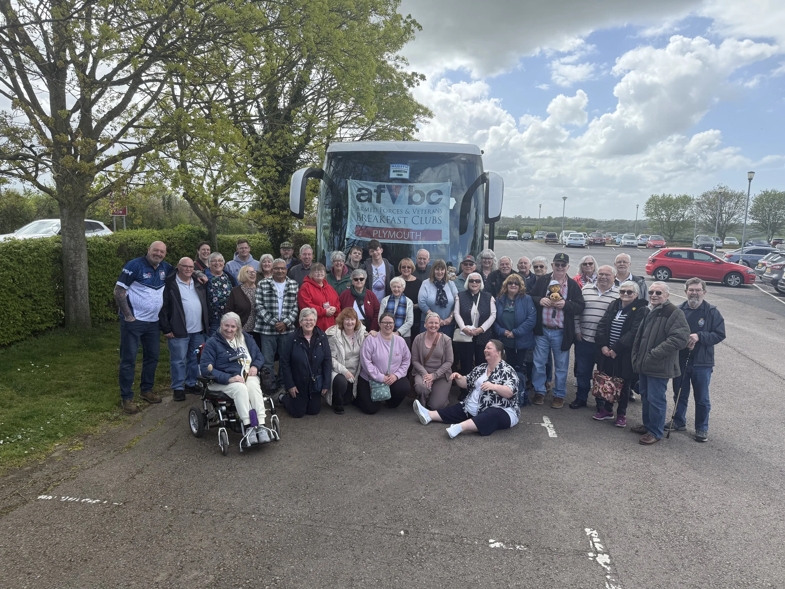Group of diverse people of all ages posing in a parking lot with a large bus behind them, trees and cloudy sky in background. Some people are smiling, among them are individuals in wheelchair and holding a pet. A banner on the bus reads 'armed forces & Veterans breakfast clubs, Plymouth.'