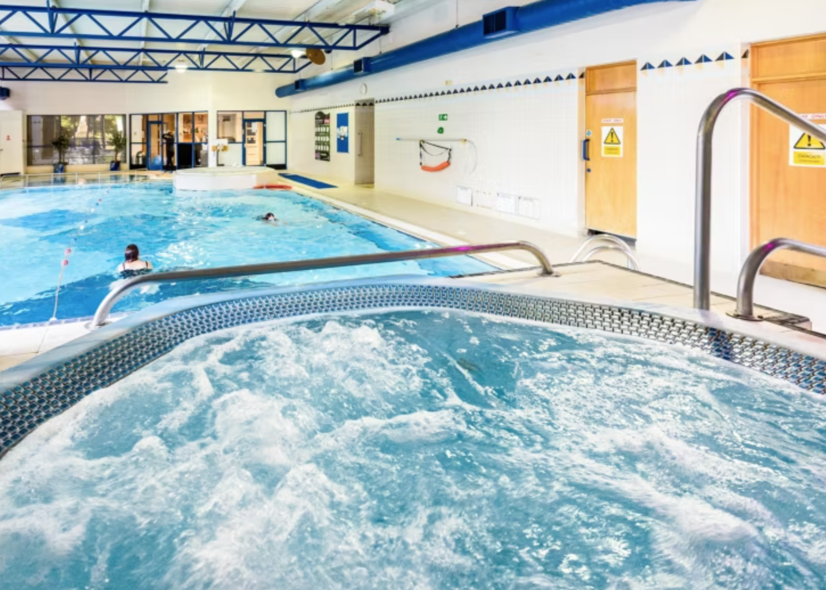 Indoor swimming pool area with a hot tub in the foreground, and a large pool with people swimming in the background. The space features blue and white color accents and safety signage.
