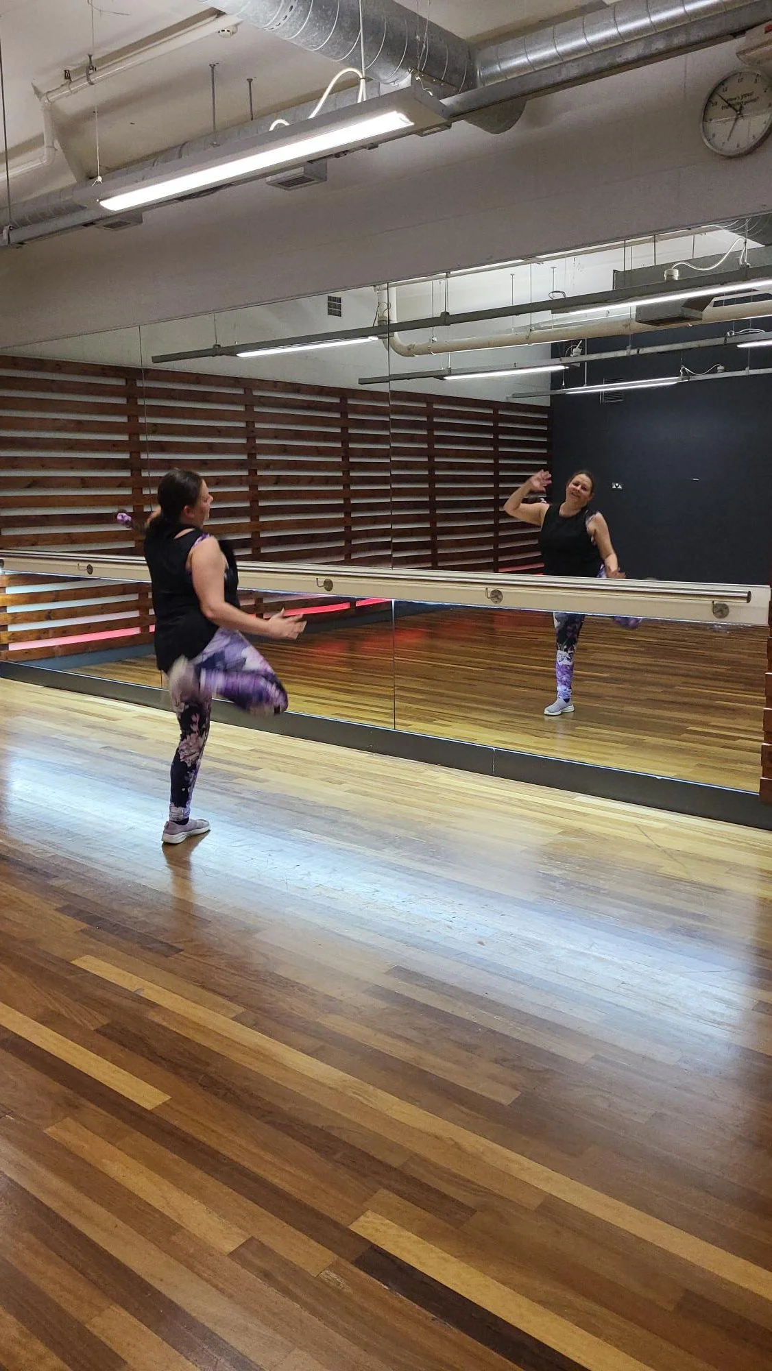 Woman in black workout top and purple patterned leggings practicing a dance move in front of a mirror in a fitness studio with wooden floors and mirrored wall.
