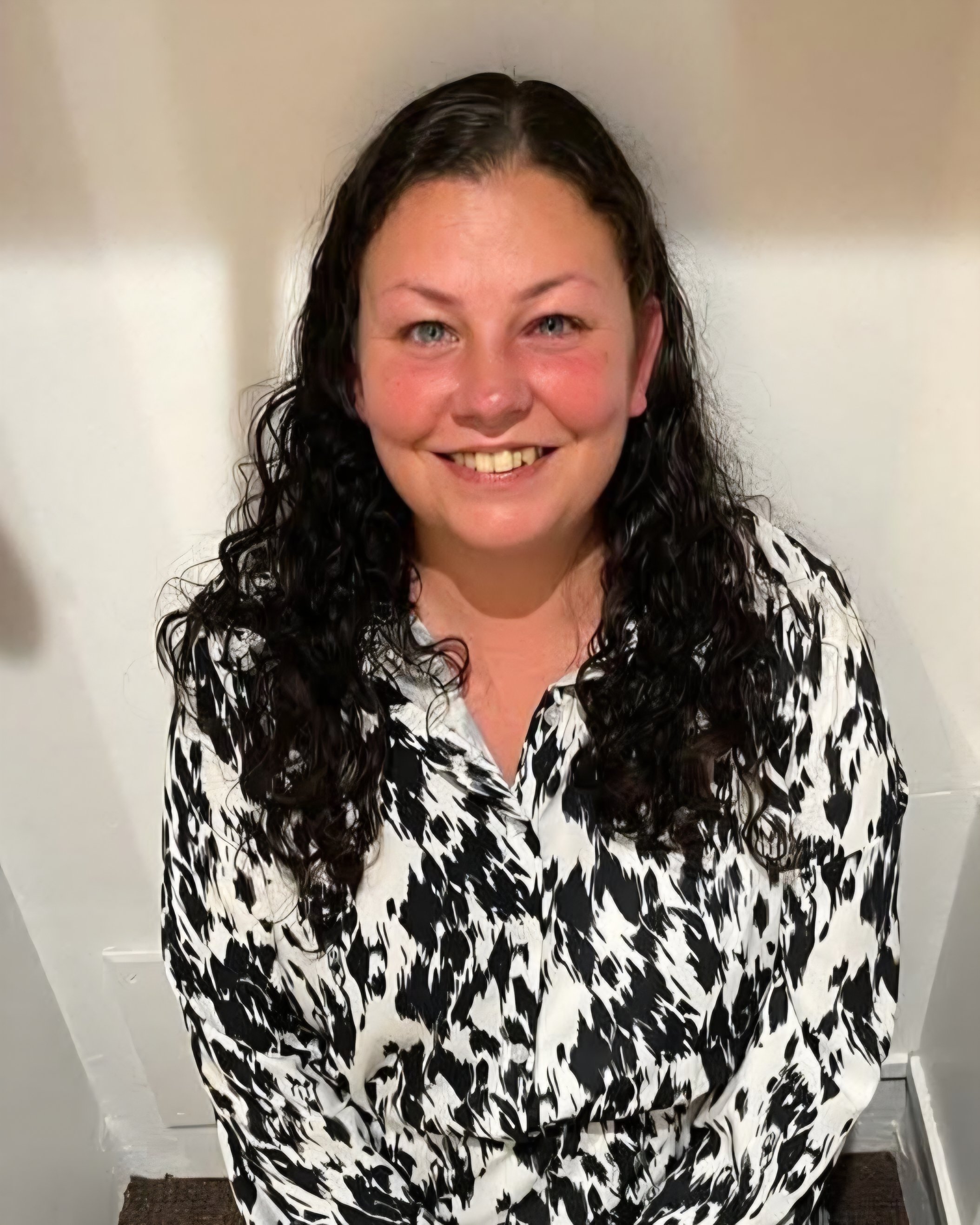 A woman with long dark curly hair and blue eyes smiling in front of a plain white wall, wearing a black and white patterned shirt.