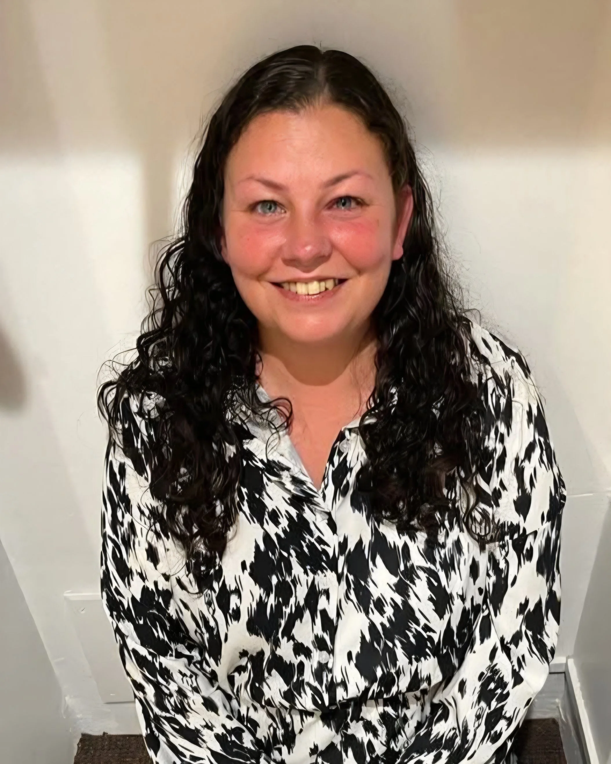 A smiling woman with wet, curly dark hair, wearing a black and white patterned top, standing in front of a light-colored wall.