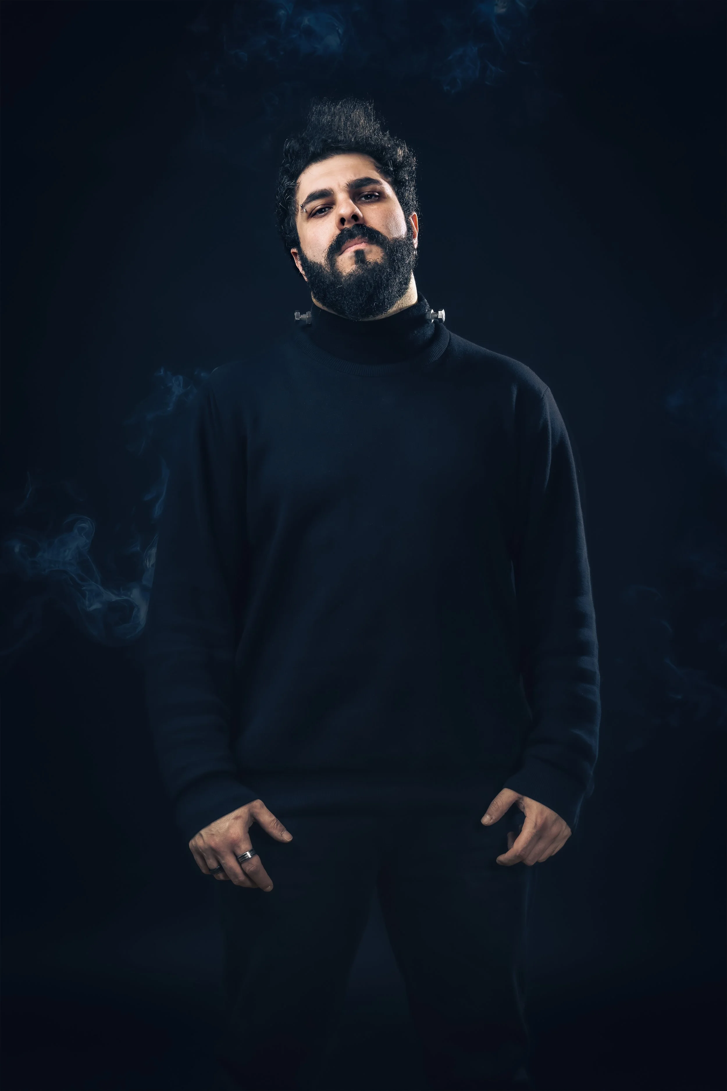 A man with dark curly hair and a full beard wearing a black long-sleeve shirt, standing against a dark background with faint smoke or mist around him, looking confidently at the camera.