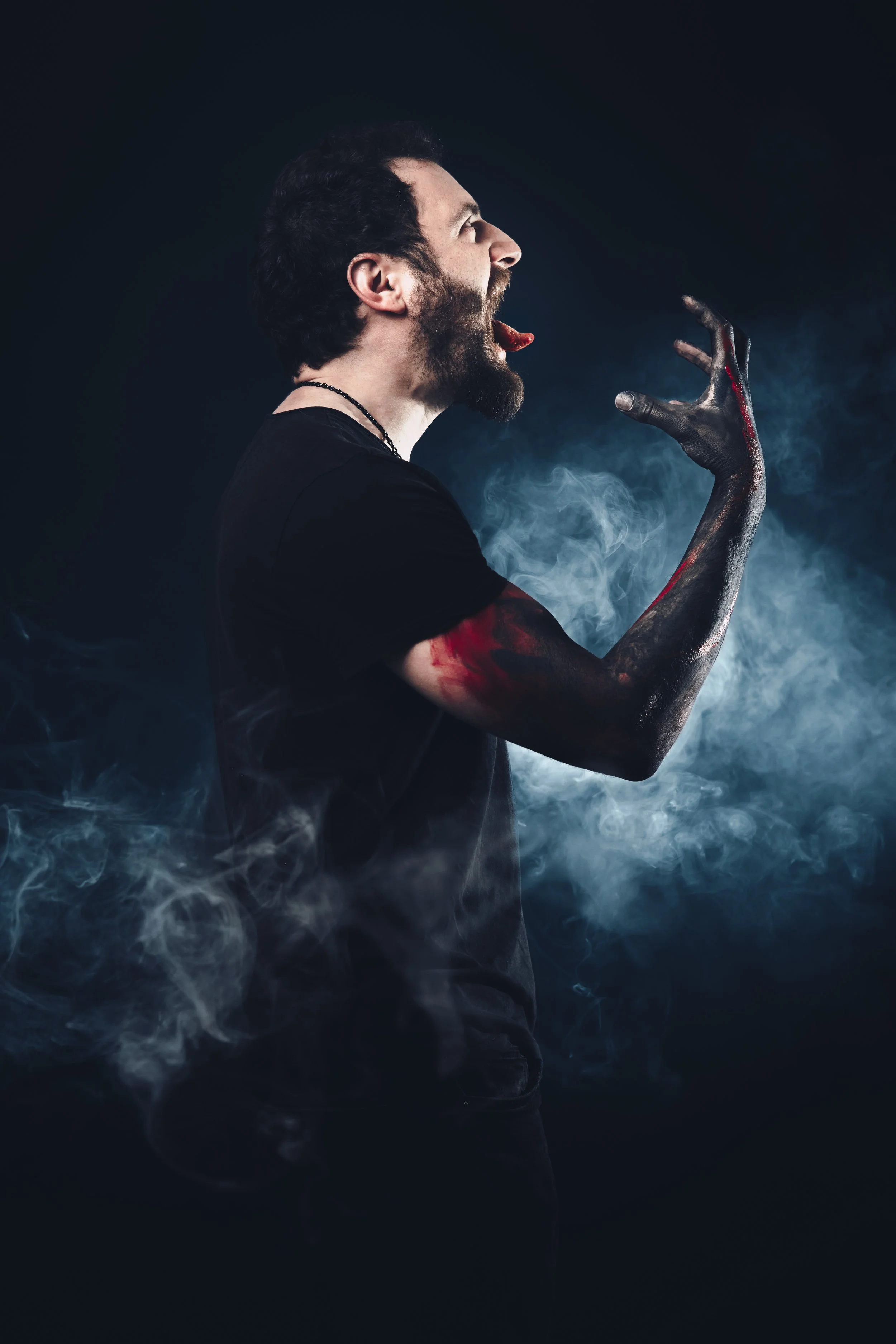 A man with a beard and dark hair is screaming, with his eyes closed and mouth wide open. He is pointing his finger as if in anger or frustration. His finger and arm are covered in dark, shiny paint or ink with some blood. The background is dark with 
