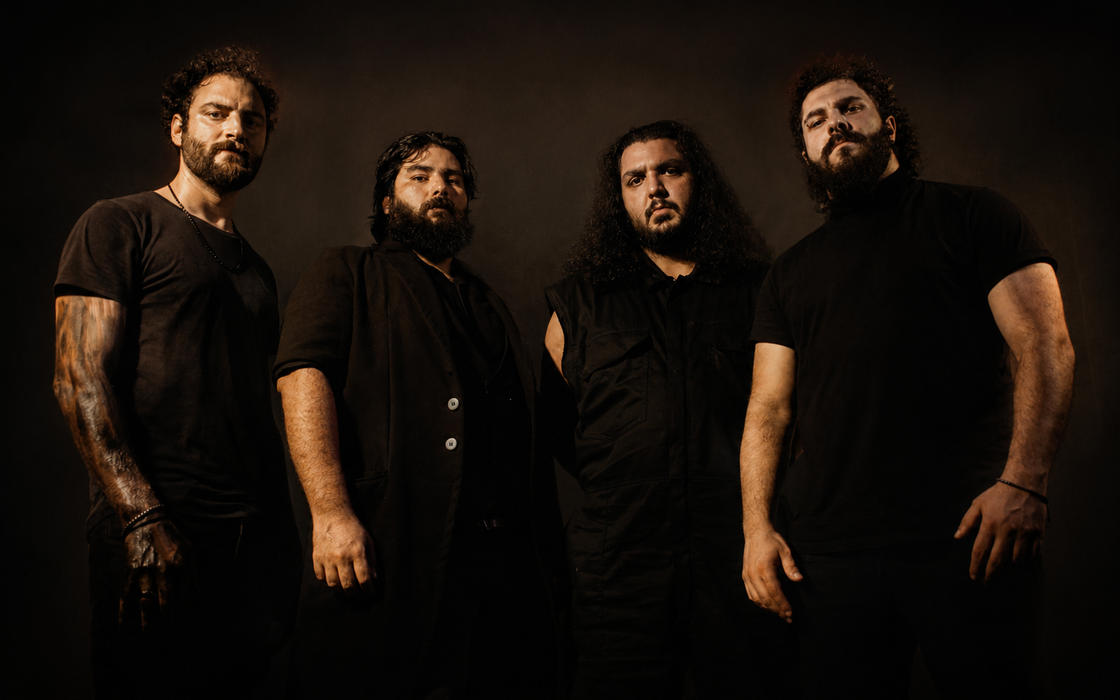 Four men with dark hair and beards, wearing black clothing, standing against a dark background.