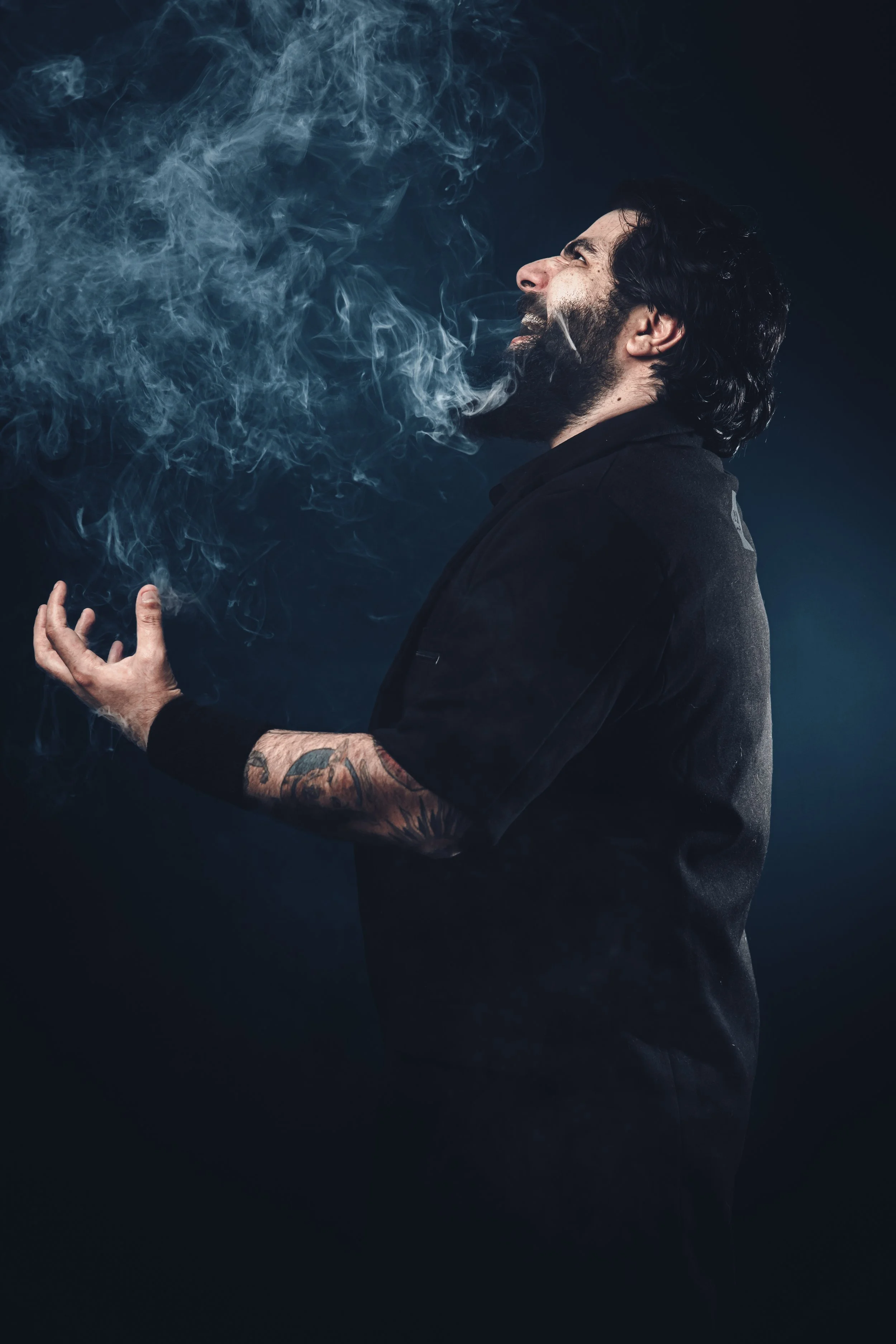 A man with dark hair and a beard, wearing a black shirt, is exhaling a large cloud of smoke against a dark background.