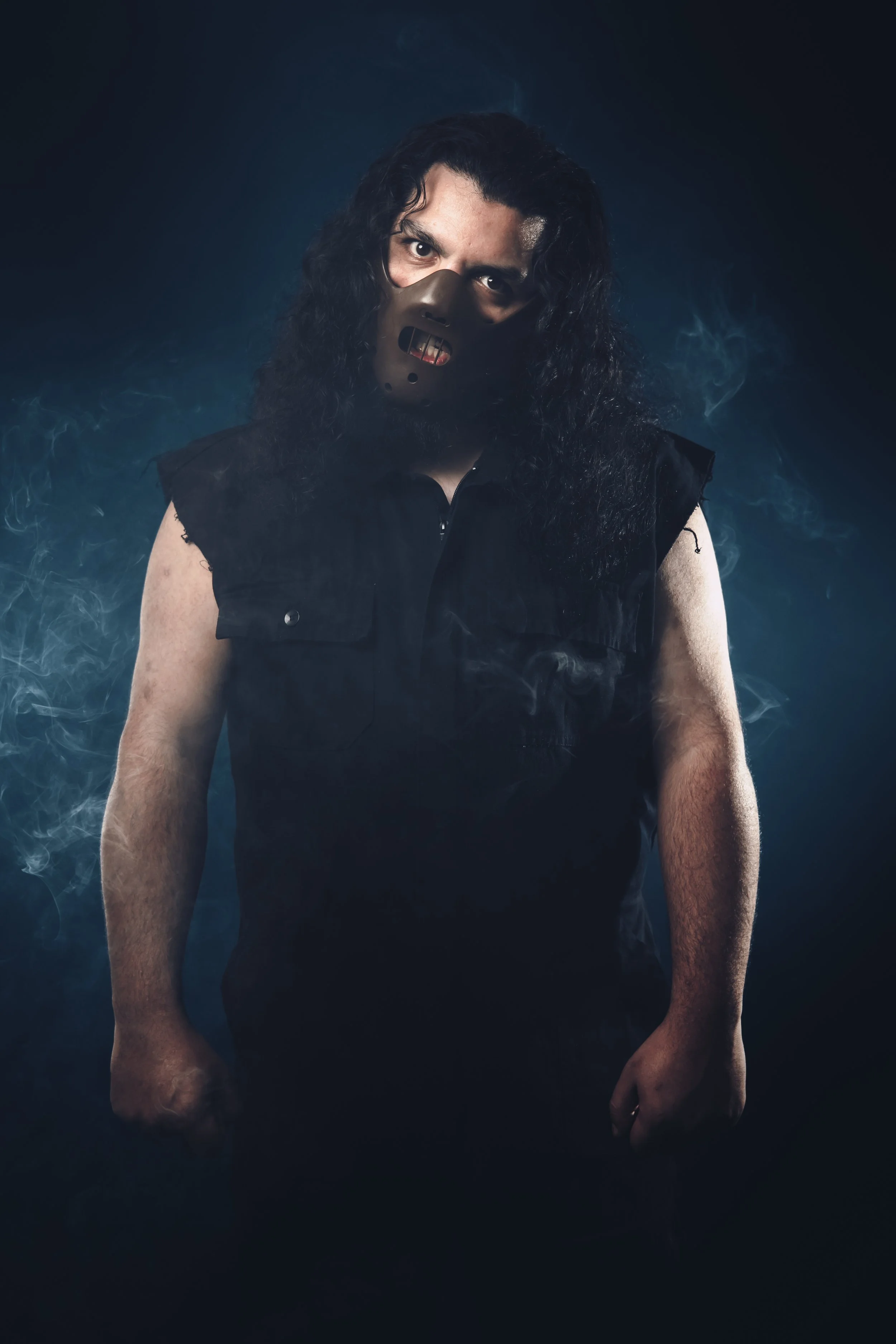 A person with long, curly black hair wearing a black sleeveless shirt and a spooky mask with gold teeth, standing against a dark background with smoky blue light.