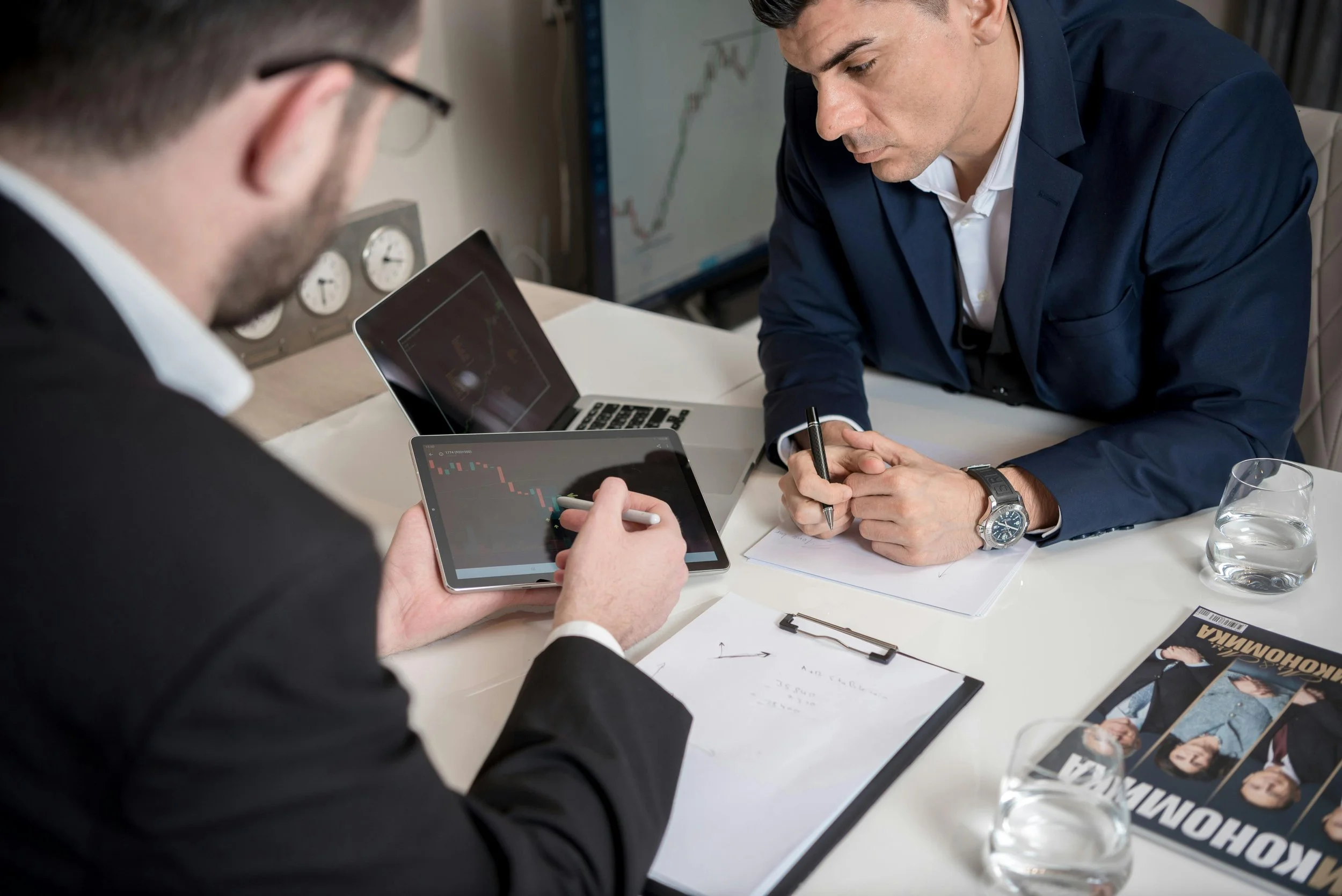 Two men in business attire analyzing stock market data during a meeting. One man is pointing at a tablet showing a trading chart, while the other man observes and takes notes. The table has papers, brochures, and a glass of water.