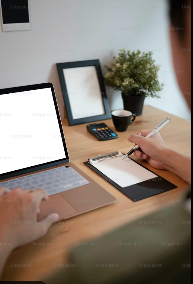 A person working at a wooden desk with a laptop, notepad, clipboard, calculator, cup of coffee, and a potted plant.