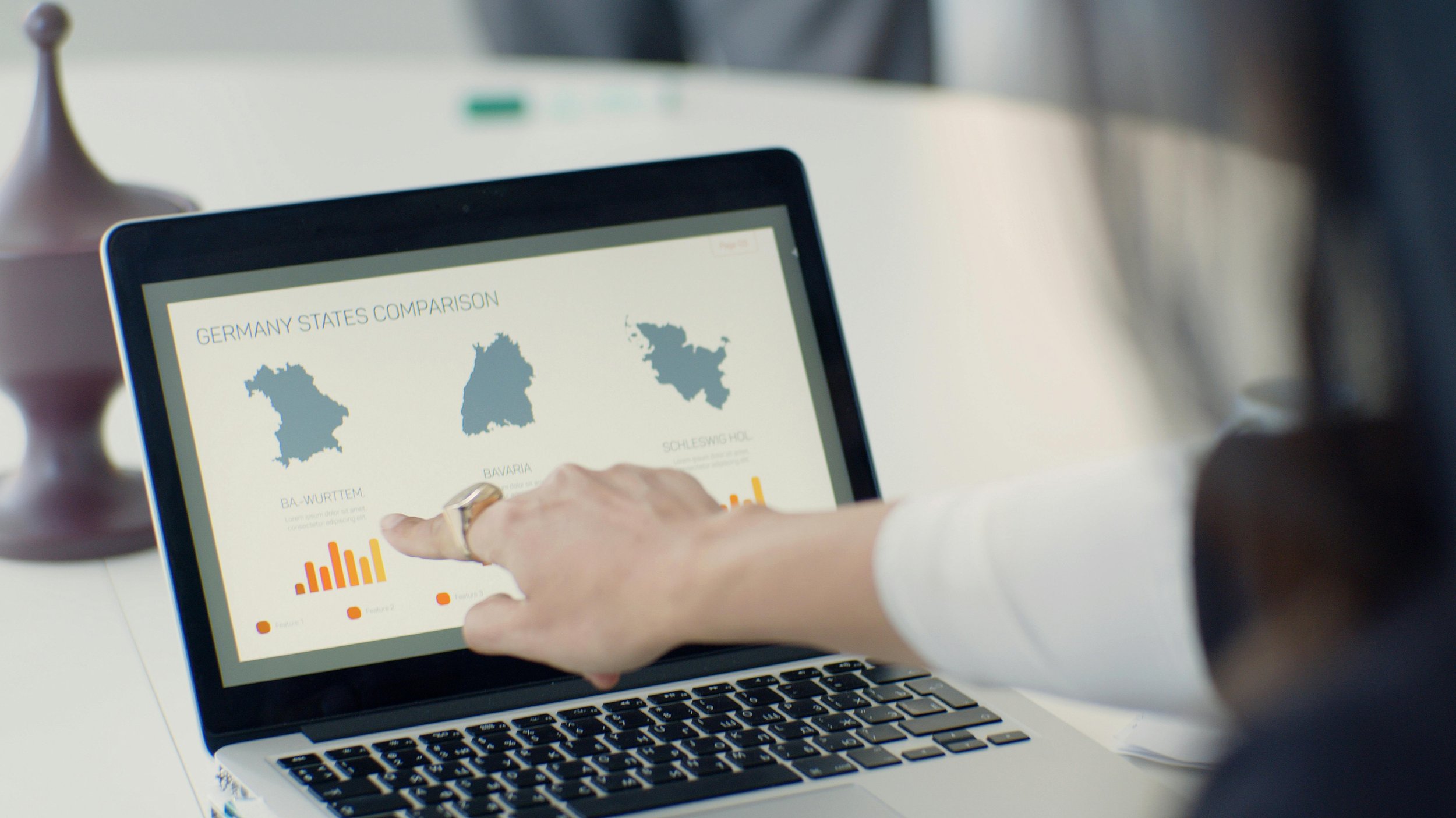 Person pointing at a tablet displaying a comparison map of Germany states, with bar graphs below, on a laptop keyboard.
