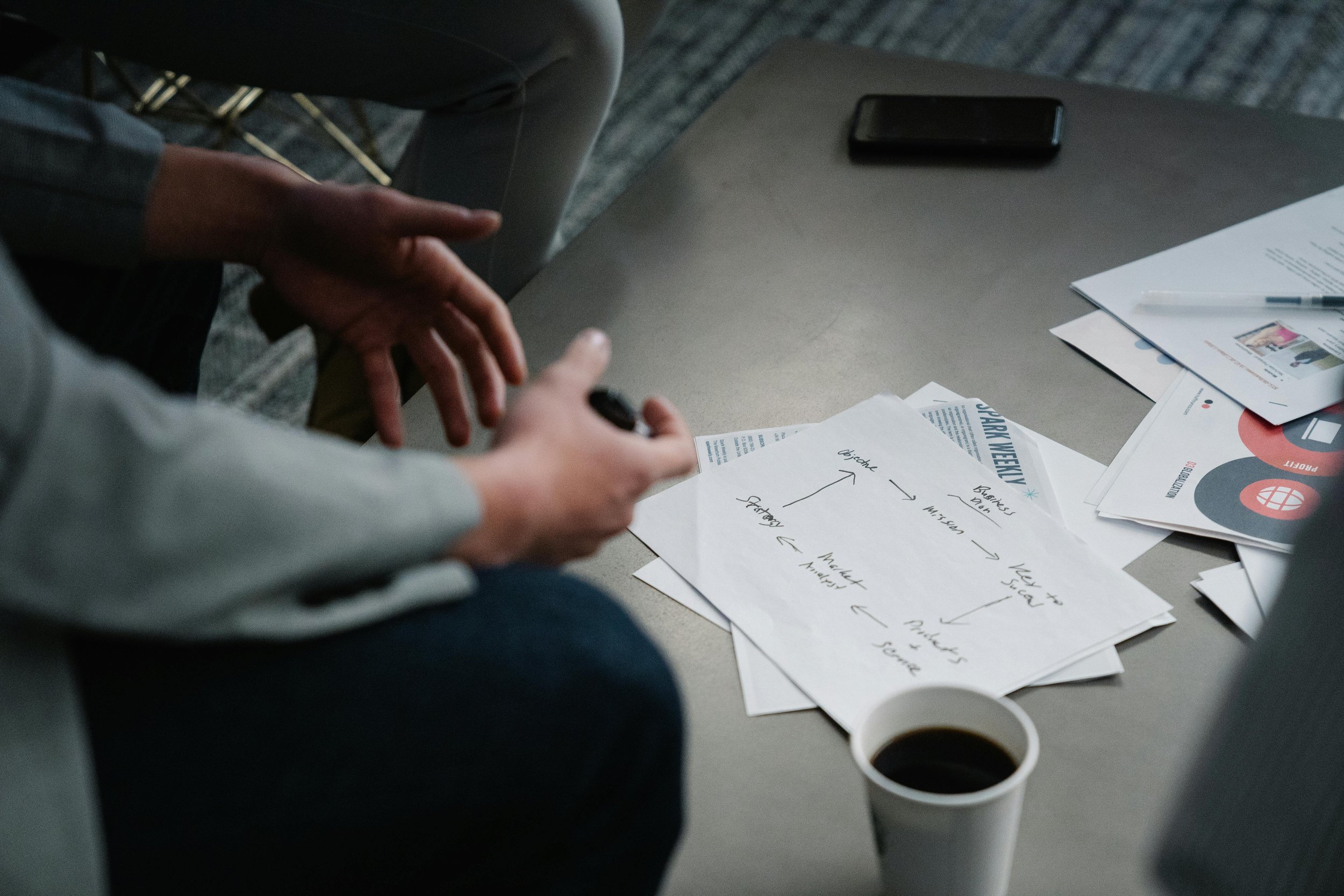 People working at a table with papers, notebooks, a pen, a smartphone, and cups of coffee.