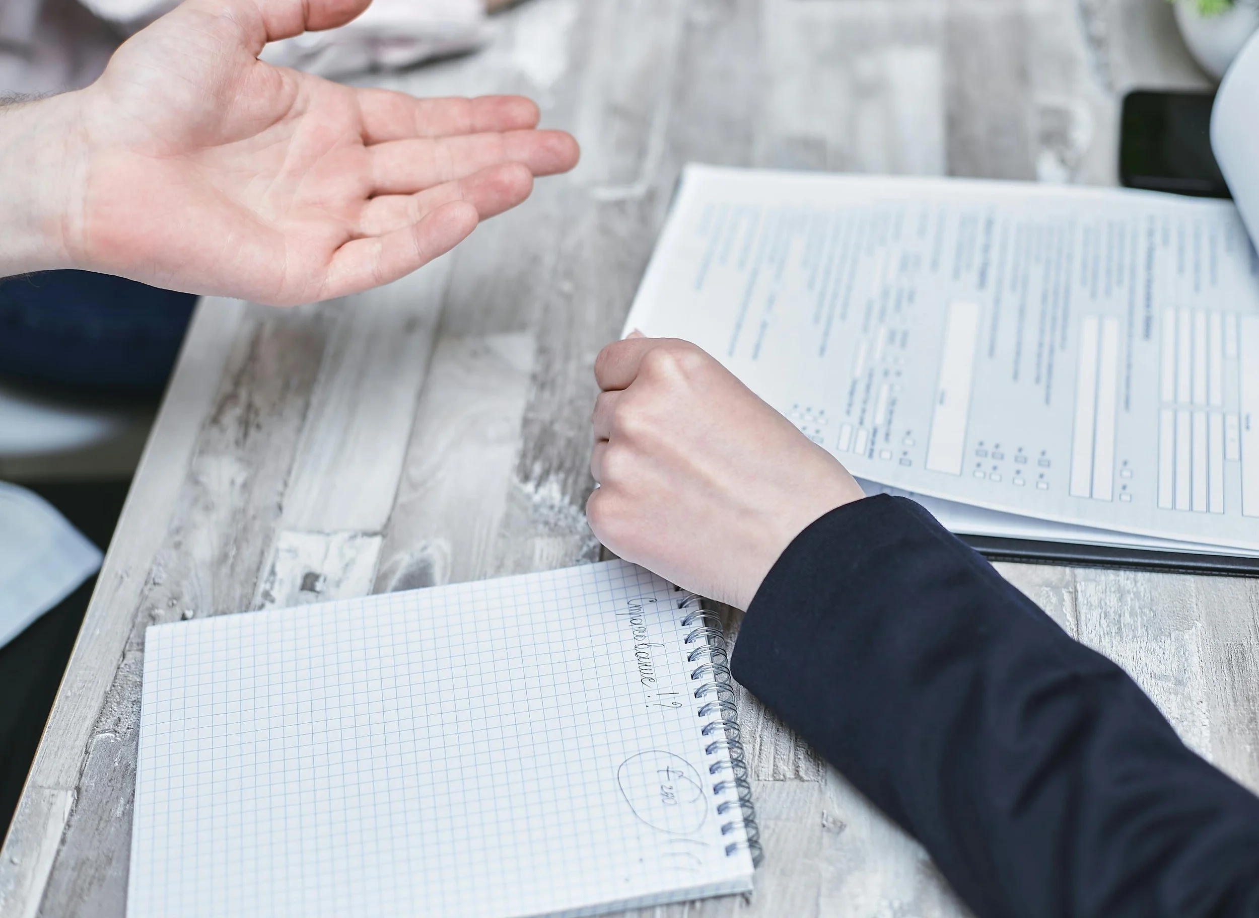 A person's hands gesturing above a notebook with grid paper on a wooden surface, with a document, a notebook, and a coffee cup partially visible in the background.