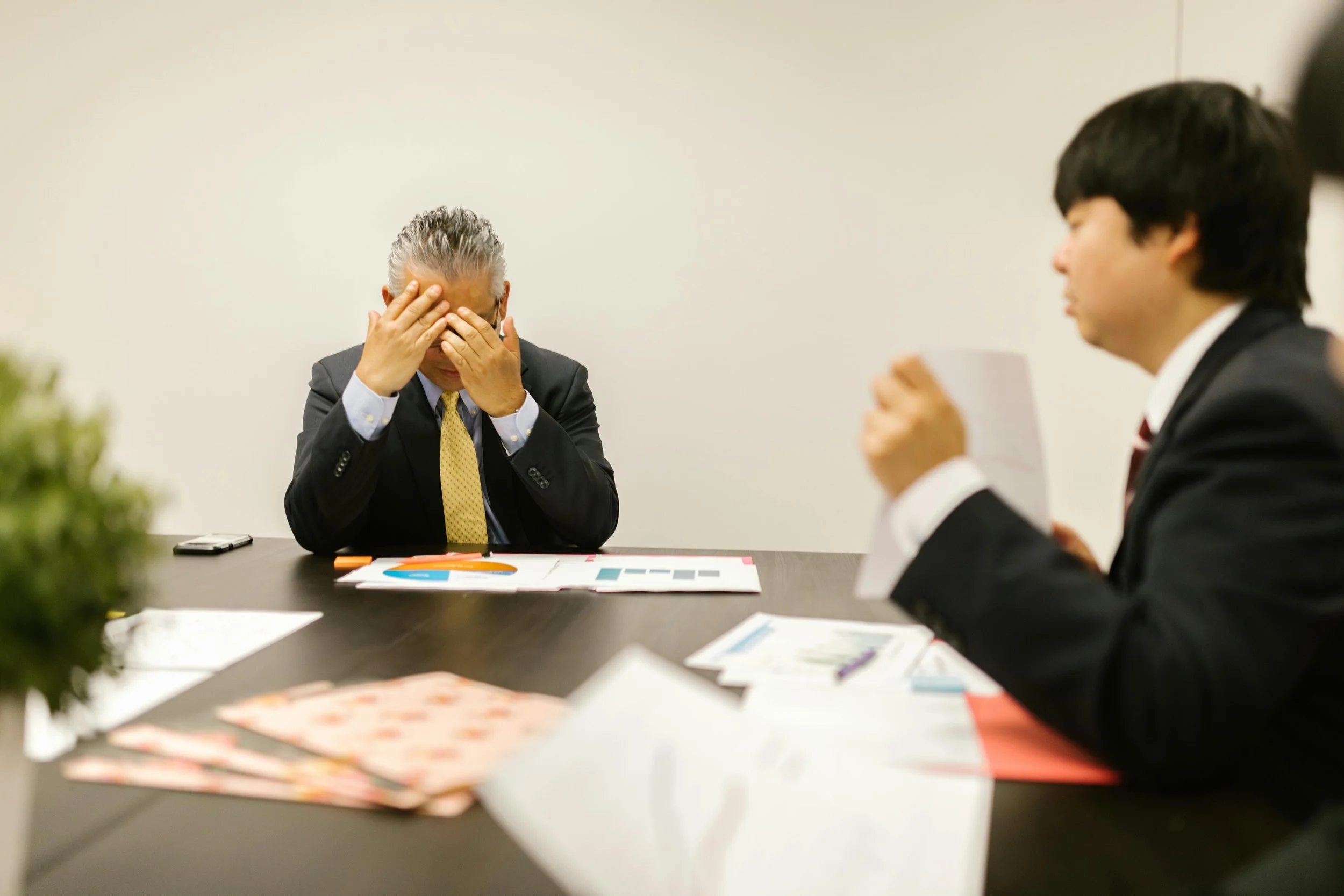 Two men in business suits are engaged in a serious discussion at a meeting table. One man, with his hair slicked back, is covering his face with his hands. The other man, with darker hair, is holding a piece of paper and speaking. The table has various documents, colorful charts, and a smartphone on it.