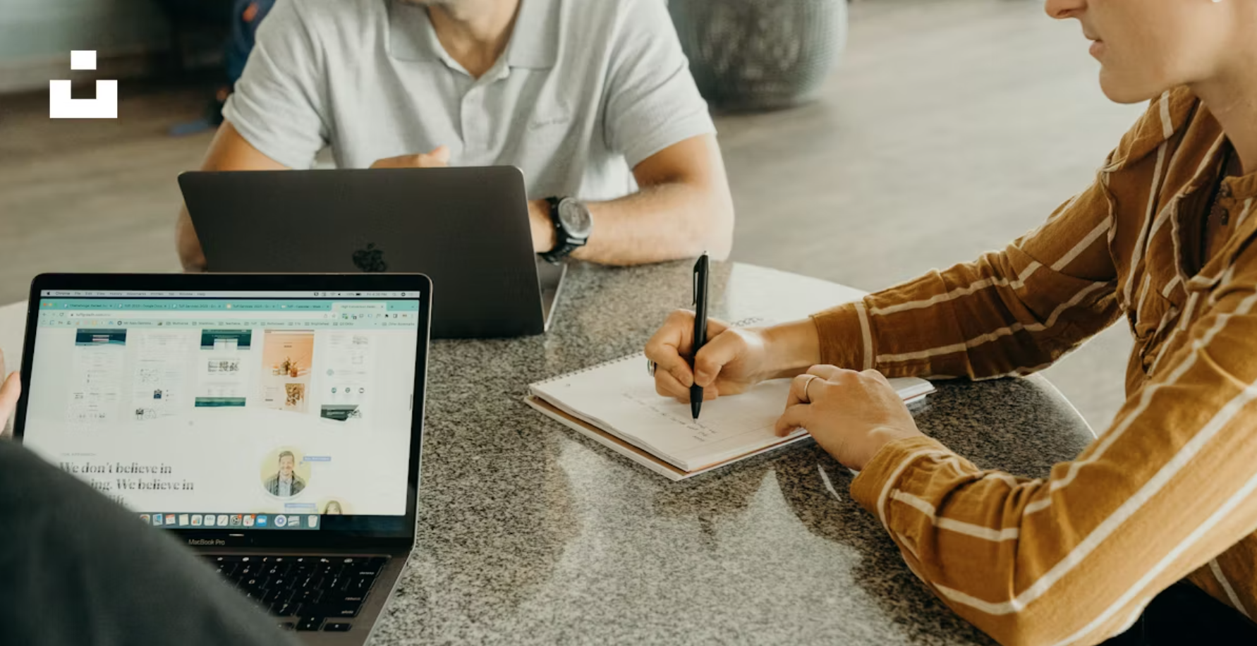 People sitting at a table working on laptops and taking notes in a casual office setting.
