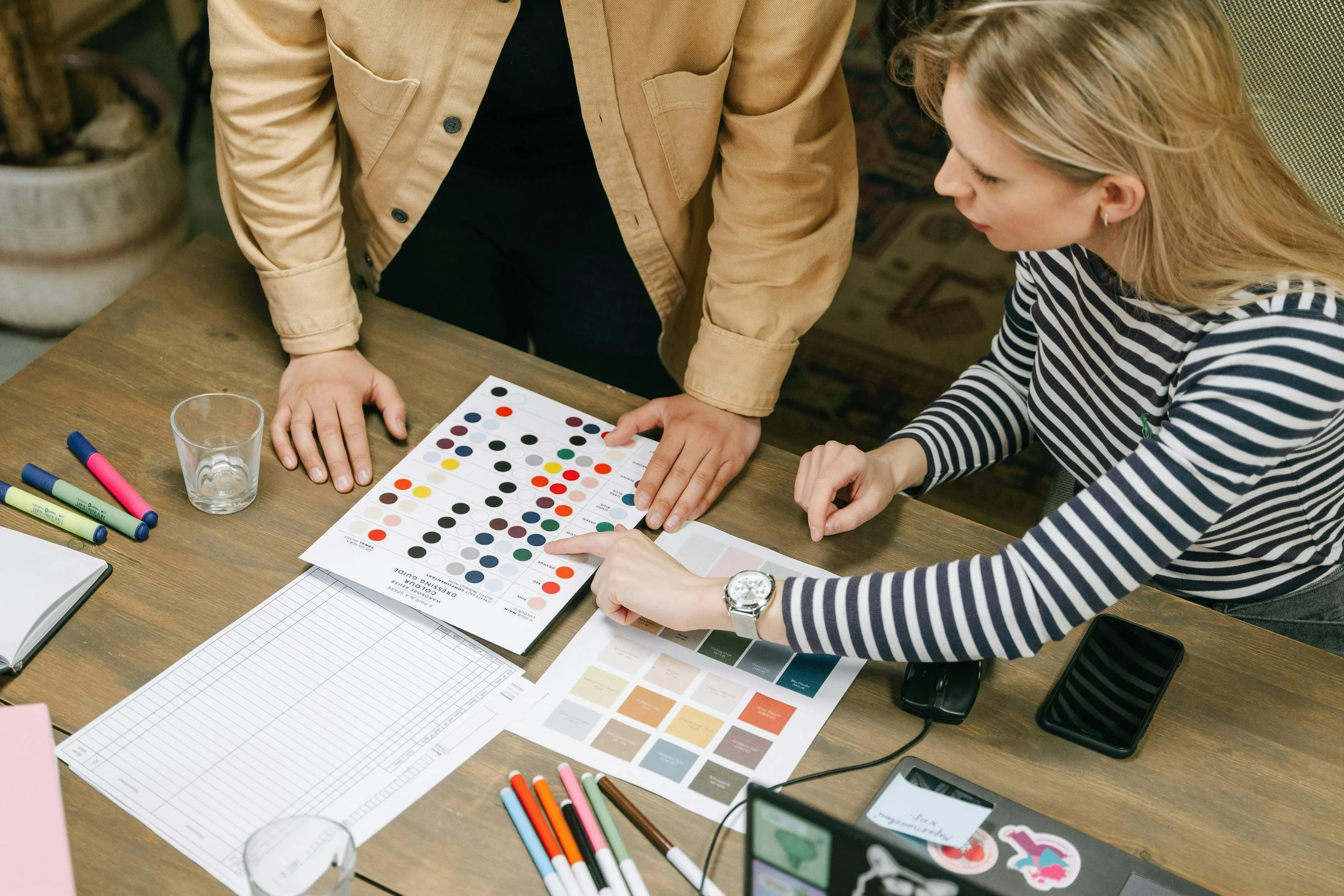 Two women are reviewing a color chart with dots on it, sitting at a cluttered table with markers, a glass, color swatches, and papers in front of them.