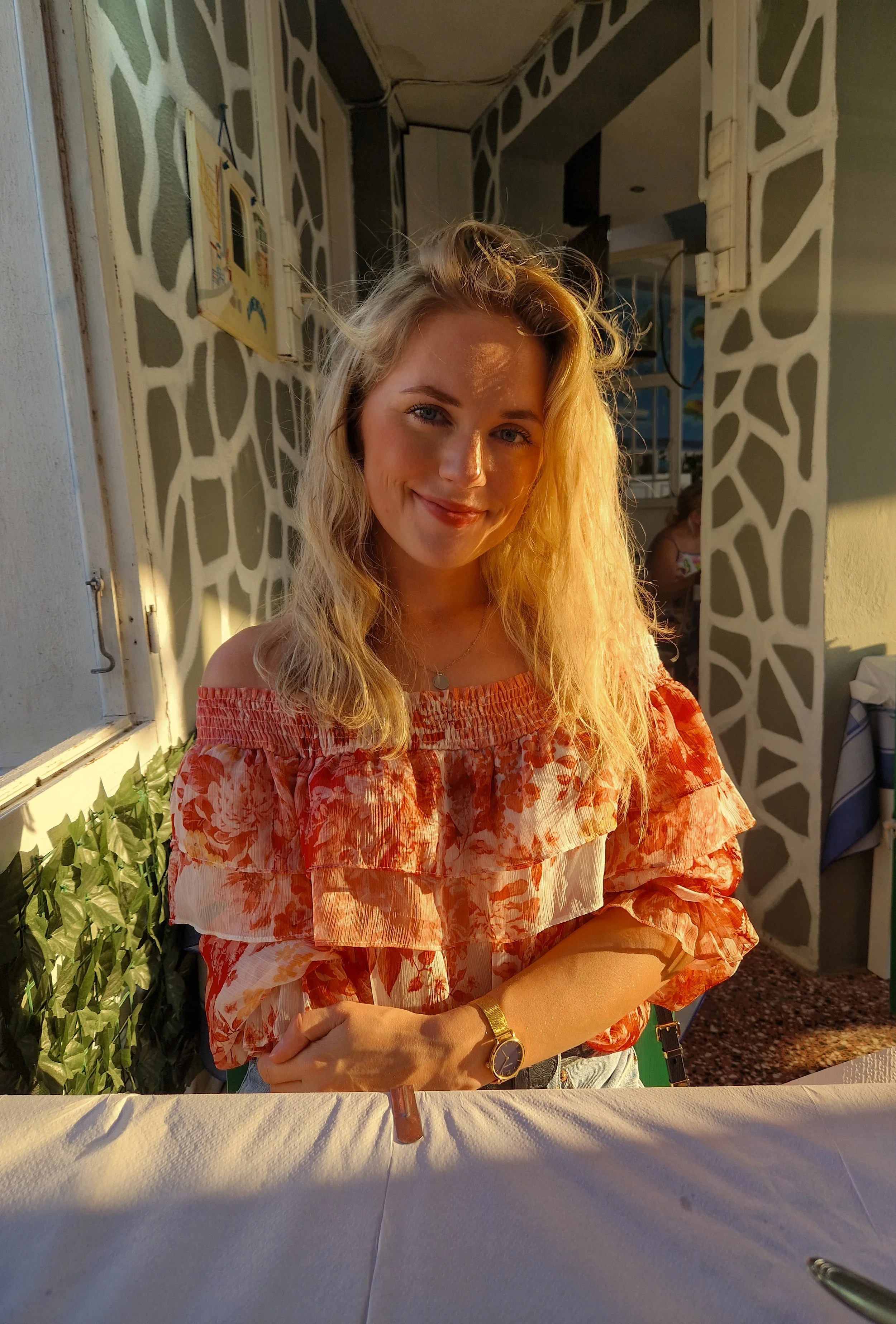 Eine junge Frau mit blonde, lockiger Haar trägt ein rotes, floral bedrucktes Oberteil. Sie steht vor einem weiß-blauen Musterhintergrund und wird von warmem Sonnenlicht beleuchtet.
