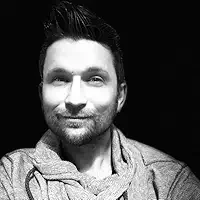 Black and white portrait of a man smiling with dark hair and a beard, wearing a light-colored shirt and scarf.