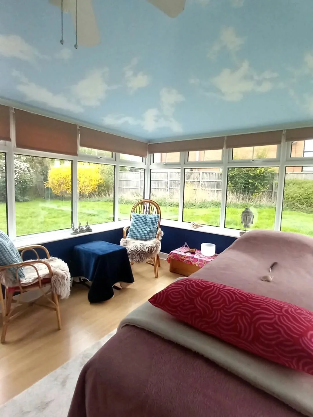 Sunroom with a sky-themed ceiling painted with clouds, large windows showing a backyard with grass and bushes, a bed with blankets and pillows, a wicker chair with a blue pillow and faux fur throw, a small table with a blue cover, and a pink patterned chest with a white lamp, overlooking the garden.