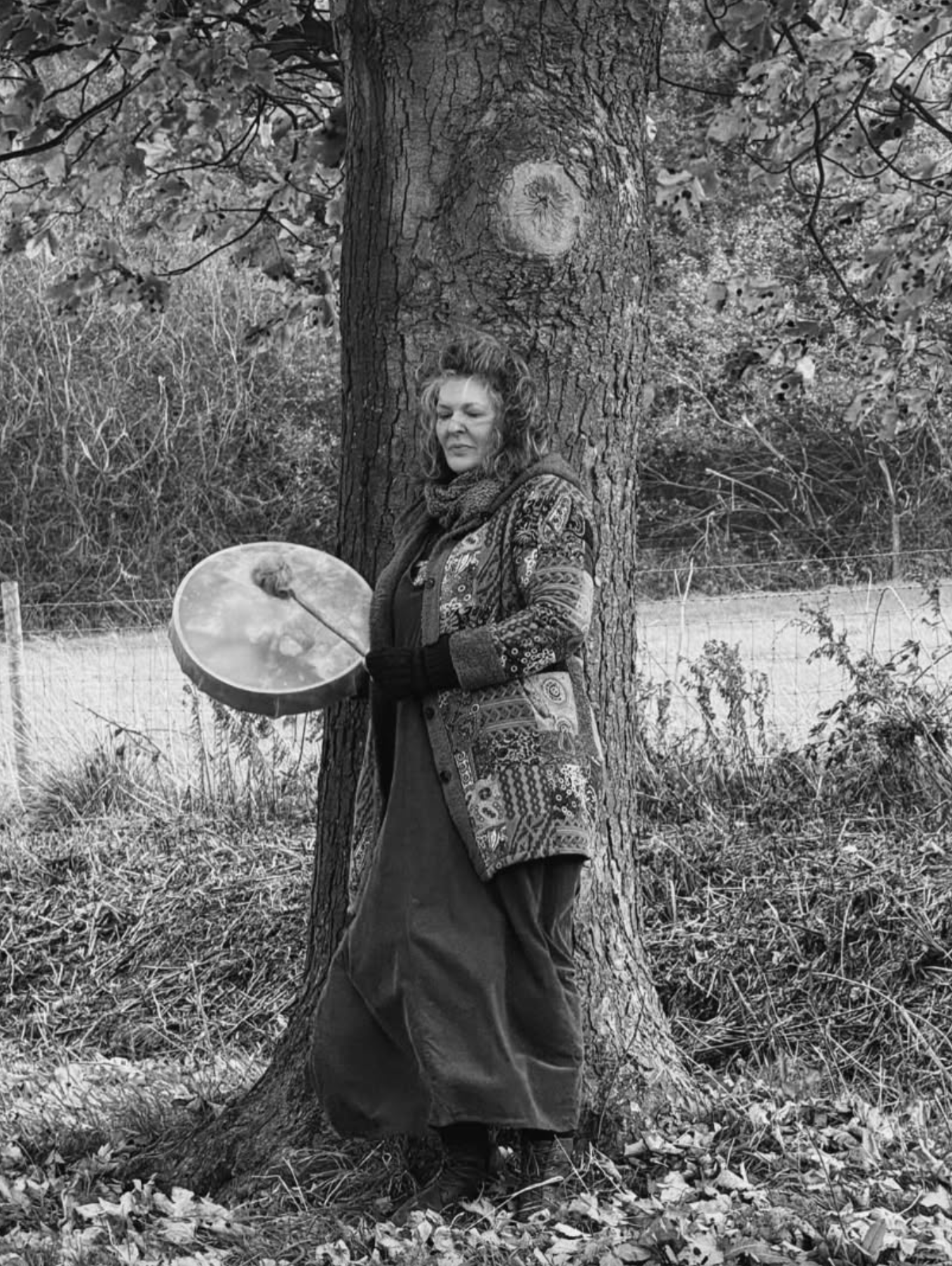 A woman standing outdoors in a wooded area, leaning against a large tree trunk while holding a frame drum with a beater in her right hand. She has curly hair and is wearing a patterned coat and a long skirt.