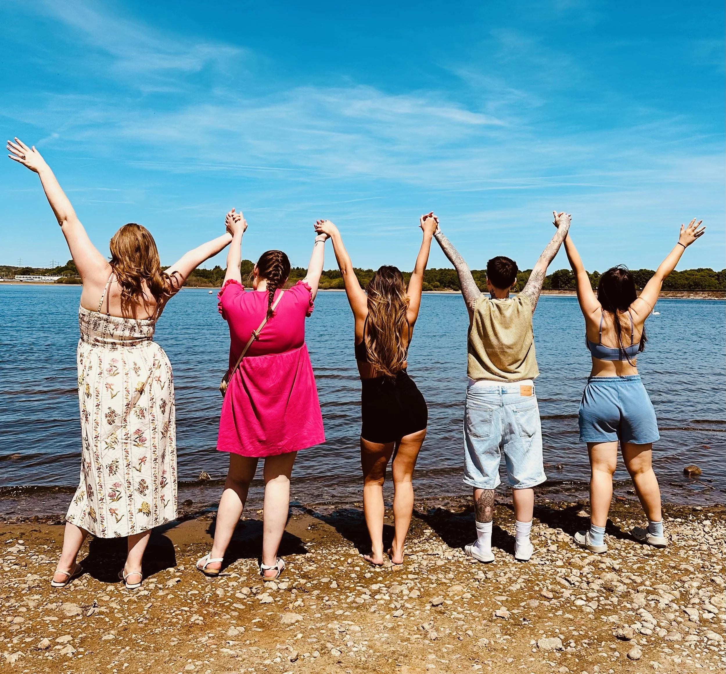 A group of people with their arms raised standing on a rocky shoreline near a body of water under a blue sky.