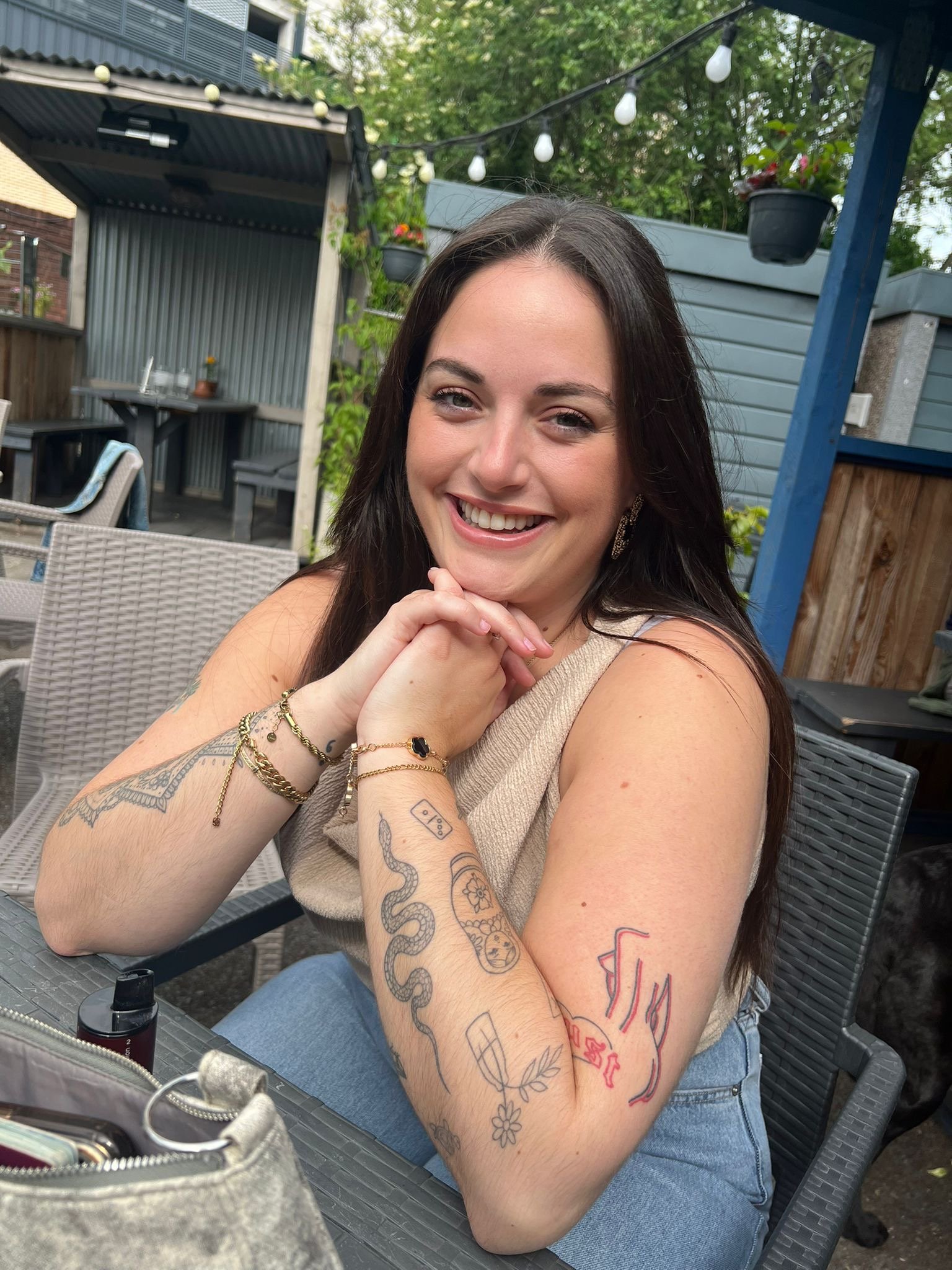 A woman with long dark hair smiling and resting her chin on her hand, sitting outdoors at a grey fence with hanging potted plants and string lights, wearing a beige top and jeans, with tattoos on her arms.