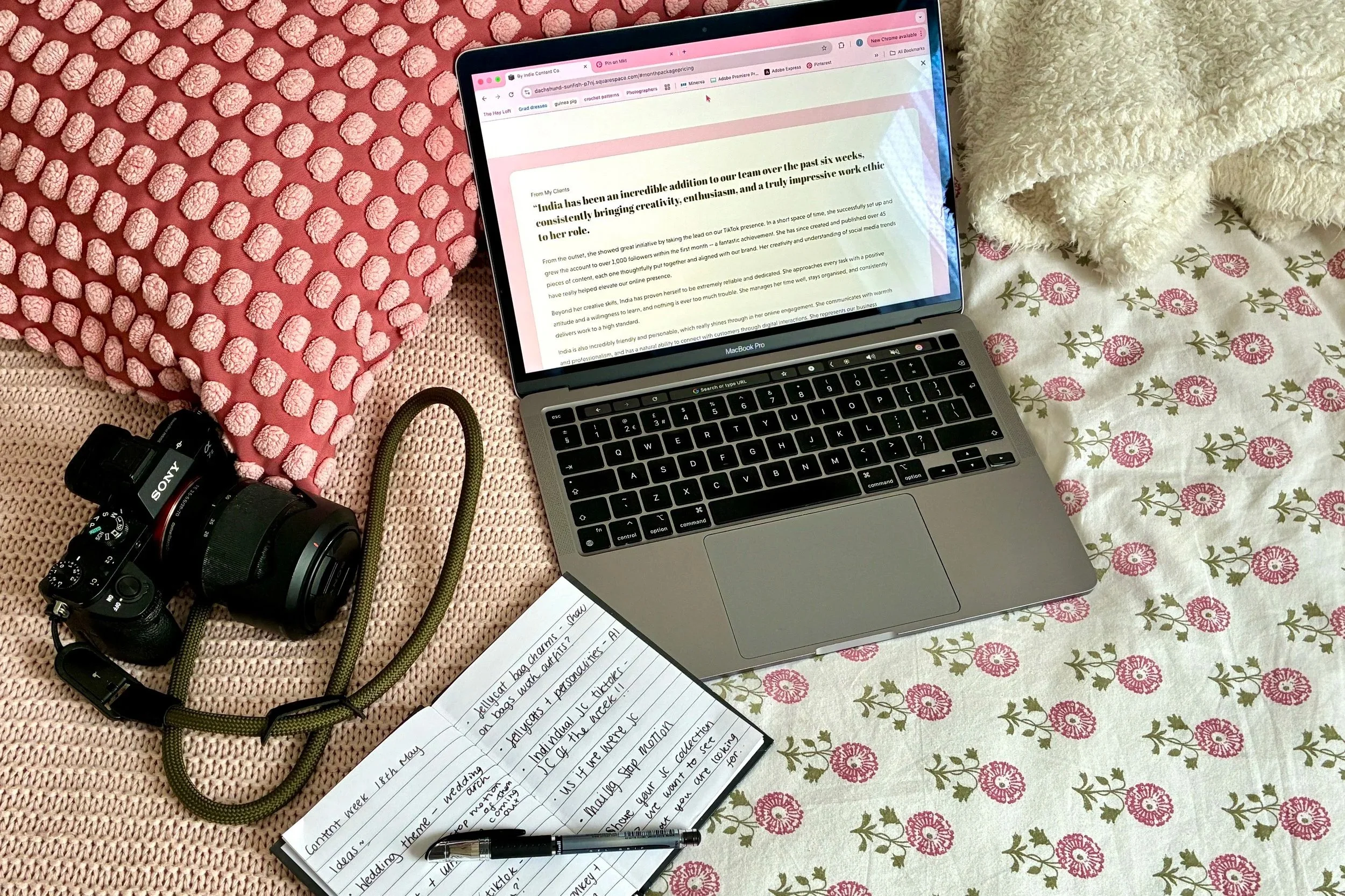 A flat lay photograph of a workspace on pink and floral bedding, featuring a camera, a green braided camera strap, an open notebook with handwritten notes, a MacBook laptop displaying a webpage, and a white plush blanket.