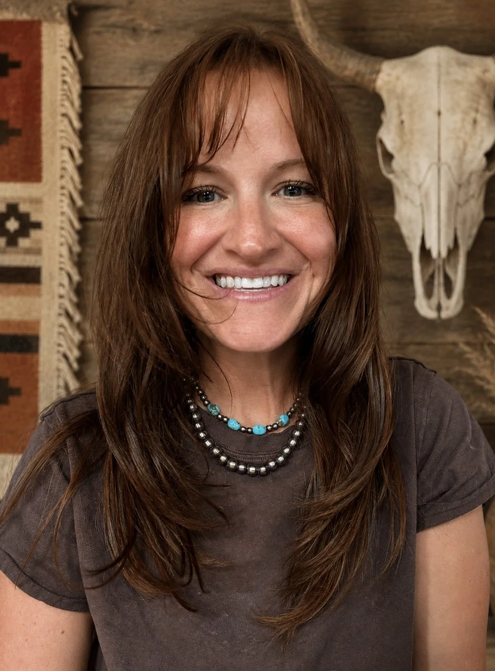 A smiling woman with long, wavy brown hair and blue eyes, wearing a dark t-shirt and layered necklaces, sitting in front of a rustic wooden wall with a mounted animal skull and a decorative woven textile.
