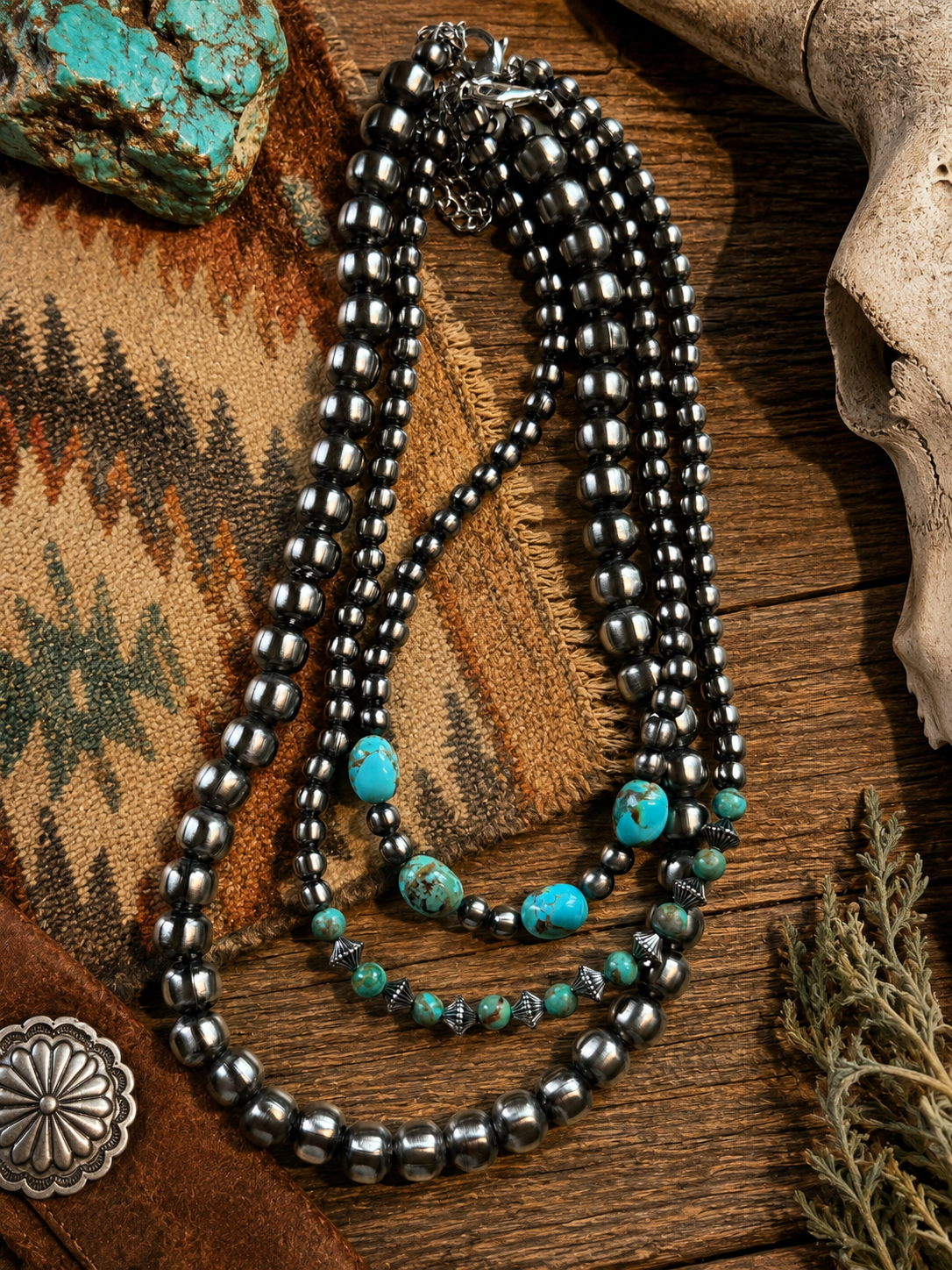 Two layered necklaces with black beads and turquoise stones on a wooden surface, surrounded by a textured fabric, a skull, a turquoise stone, a silver flower-shaped ornament, and dried plants.